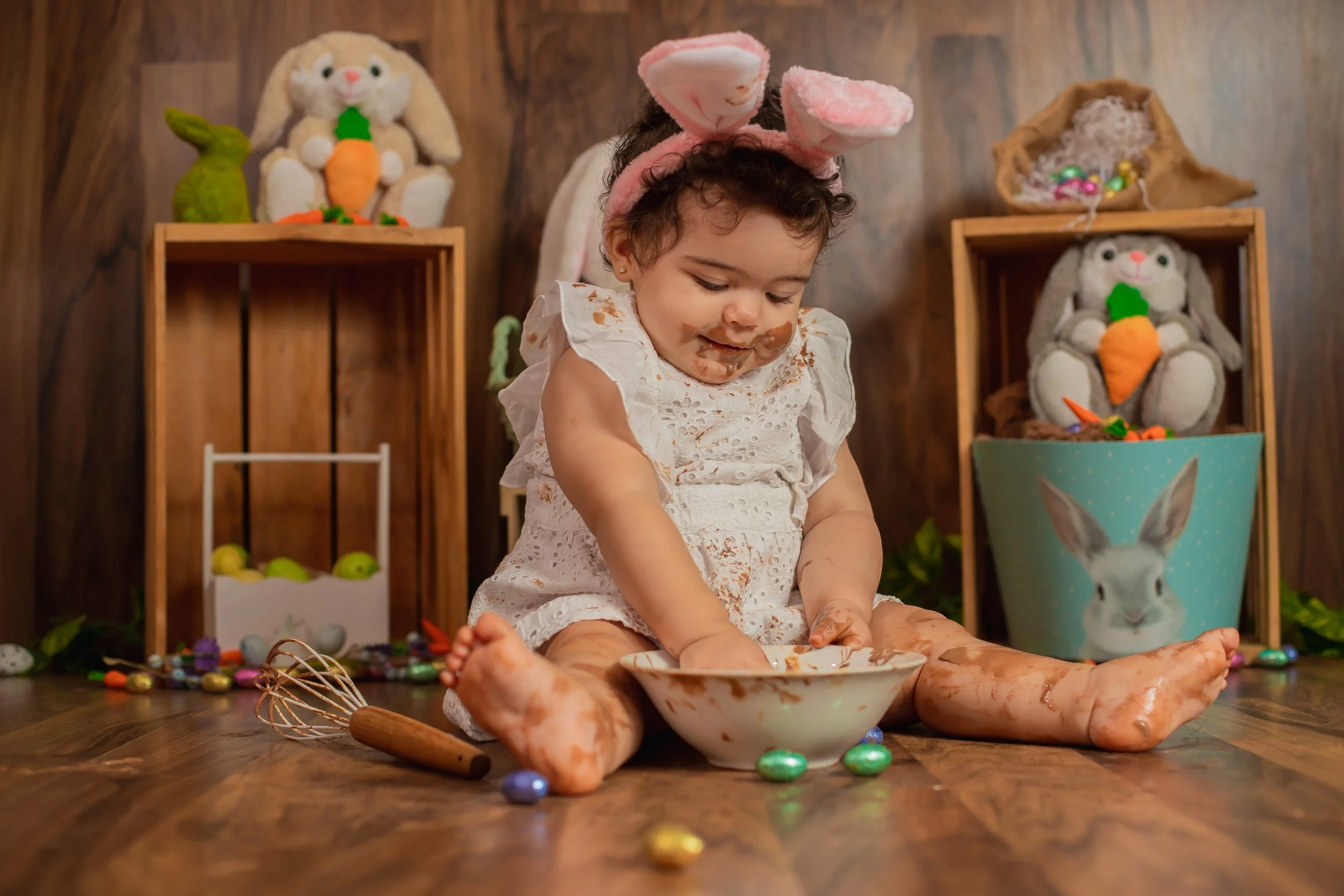 Young child dressed in white with pink bunny ears headband, sitting on a wooden floor, covered in chocolate, playing with a bowl and surrounded by colorful candies and Easter decorations in a festive setting.
