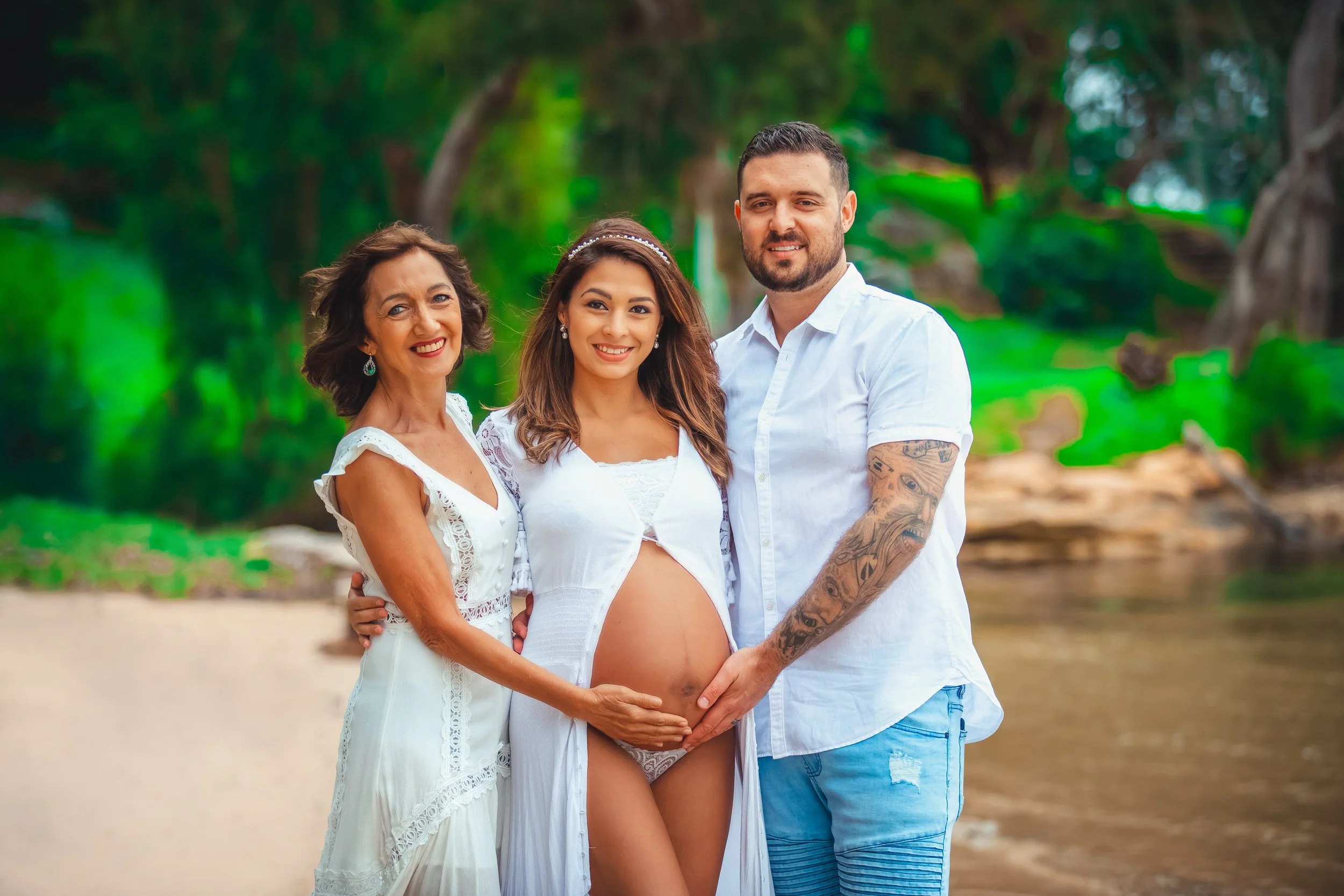 Family of three on a beach, with a pregnant woman in the middle, smiling, with her parents on either side, holding her belly.