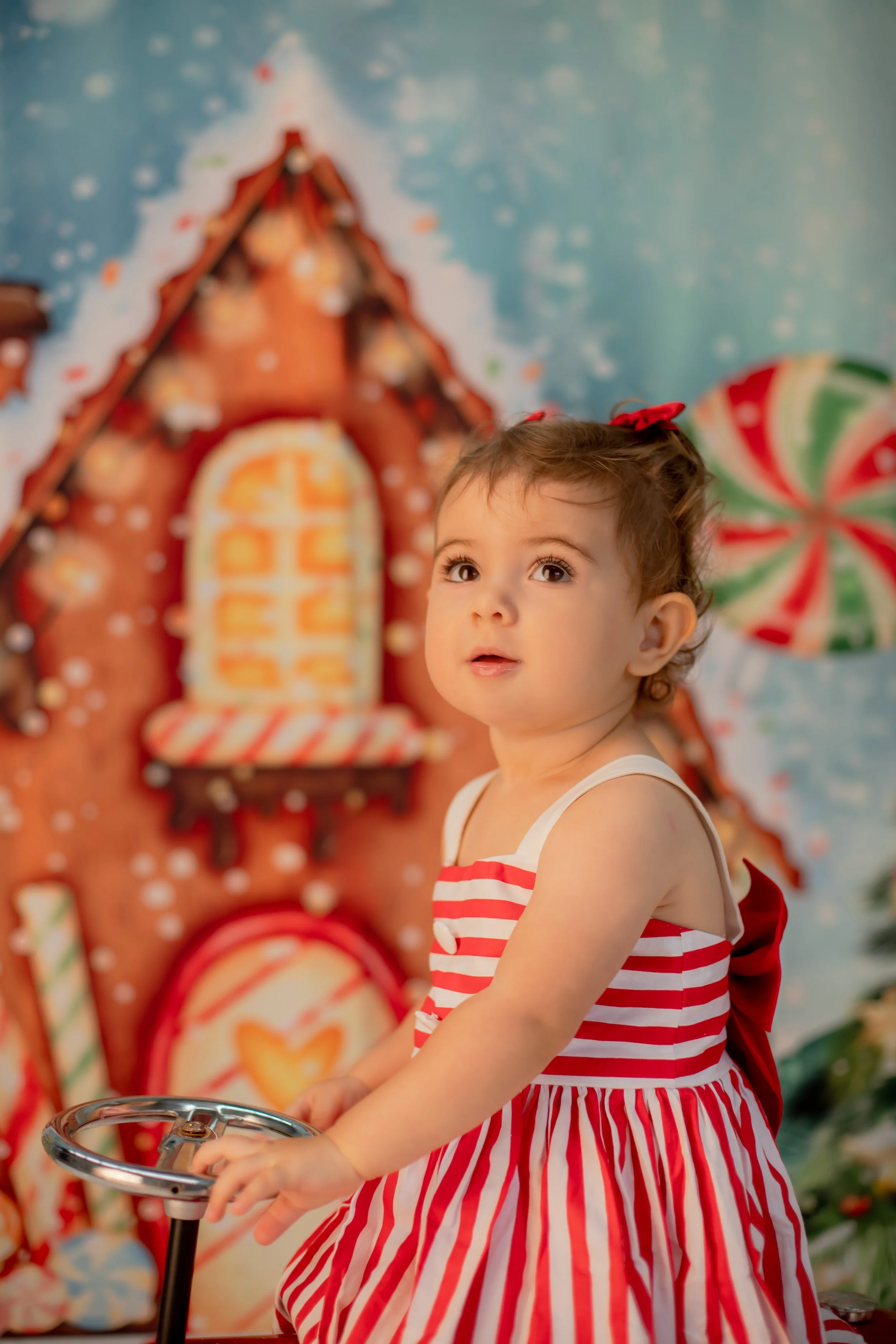 A young girl in a red and white striped dress with a red bow in her hair, riding a small tricycle in front of a festive gingerbread house and holiday decorations.