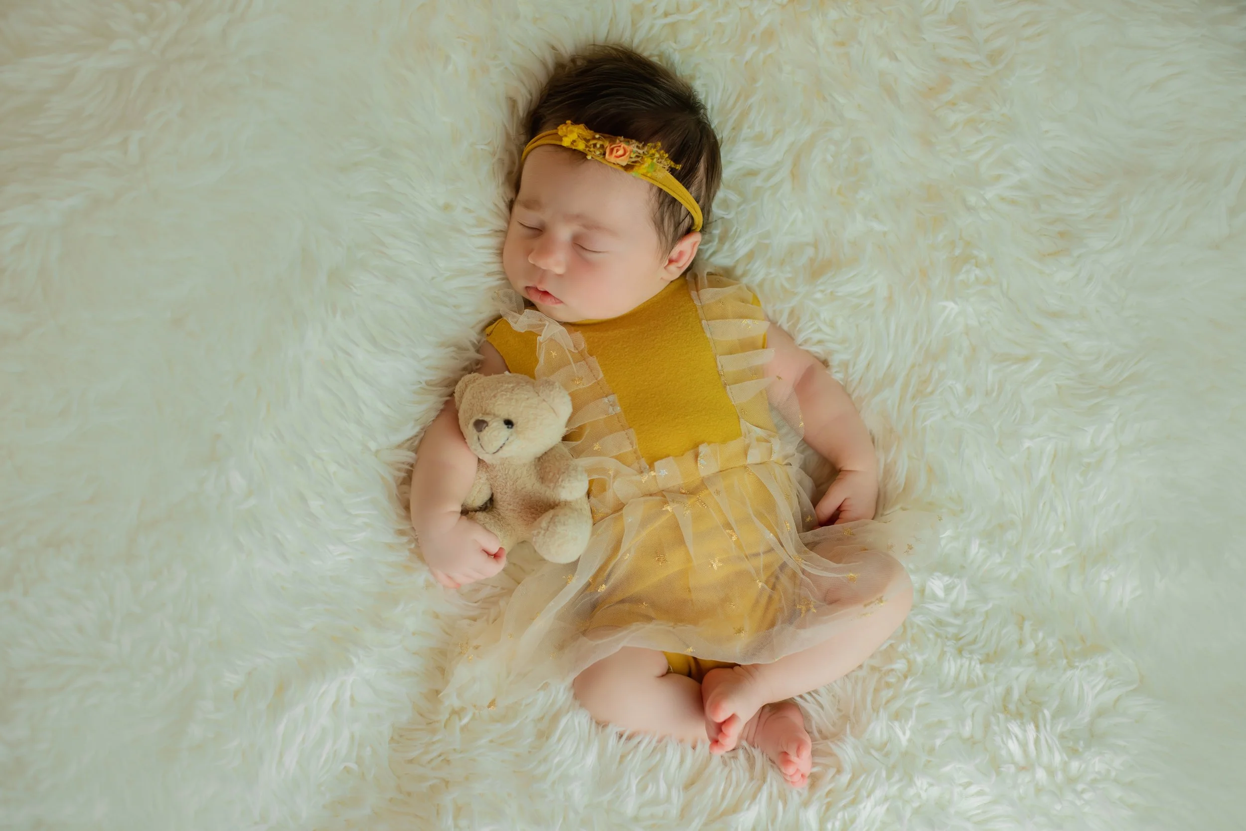 A sleeping baby girl lying on a white furry blanket, holding a small teddy bear, wearing a yellow dress and a yellow headband.