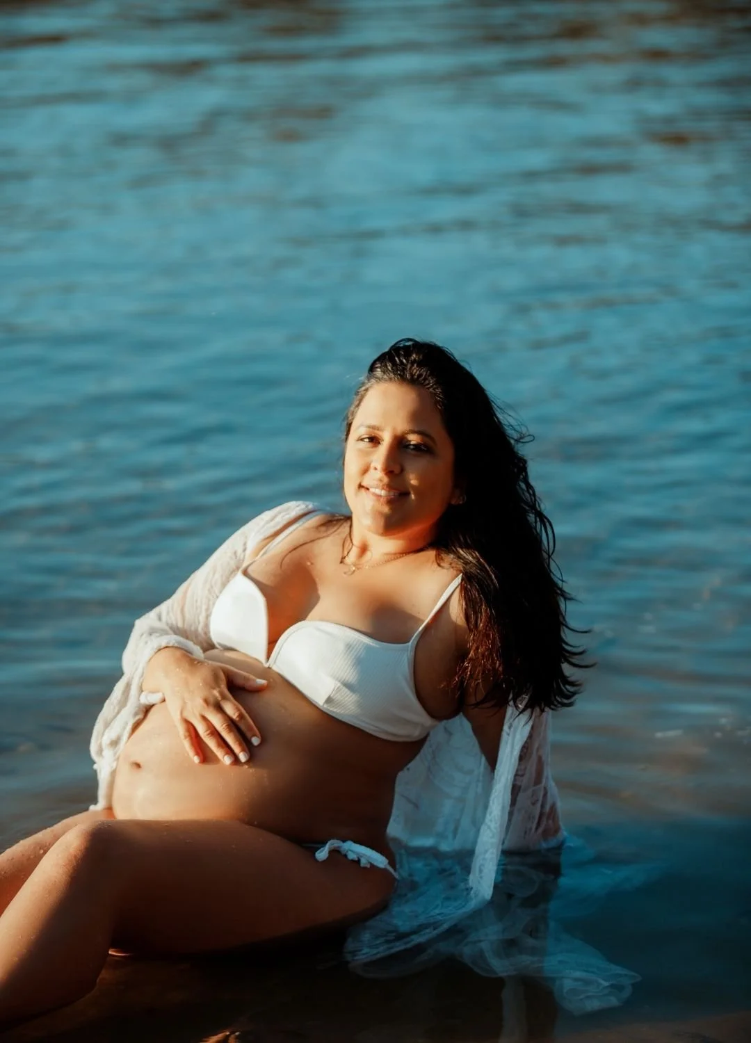 A woman in a white swimsuit and cover-up sitting in the water at the beach, with sunlight illuminating her face and hair, and a calm ocean in the background.