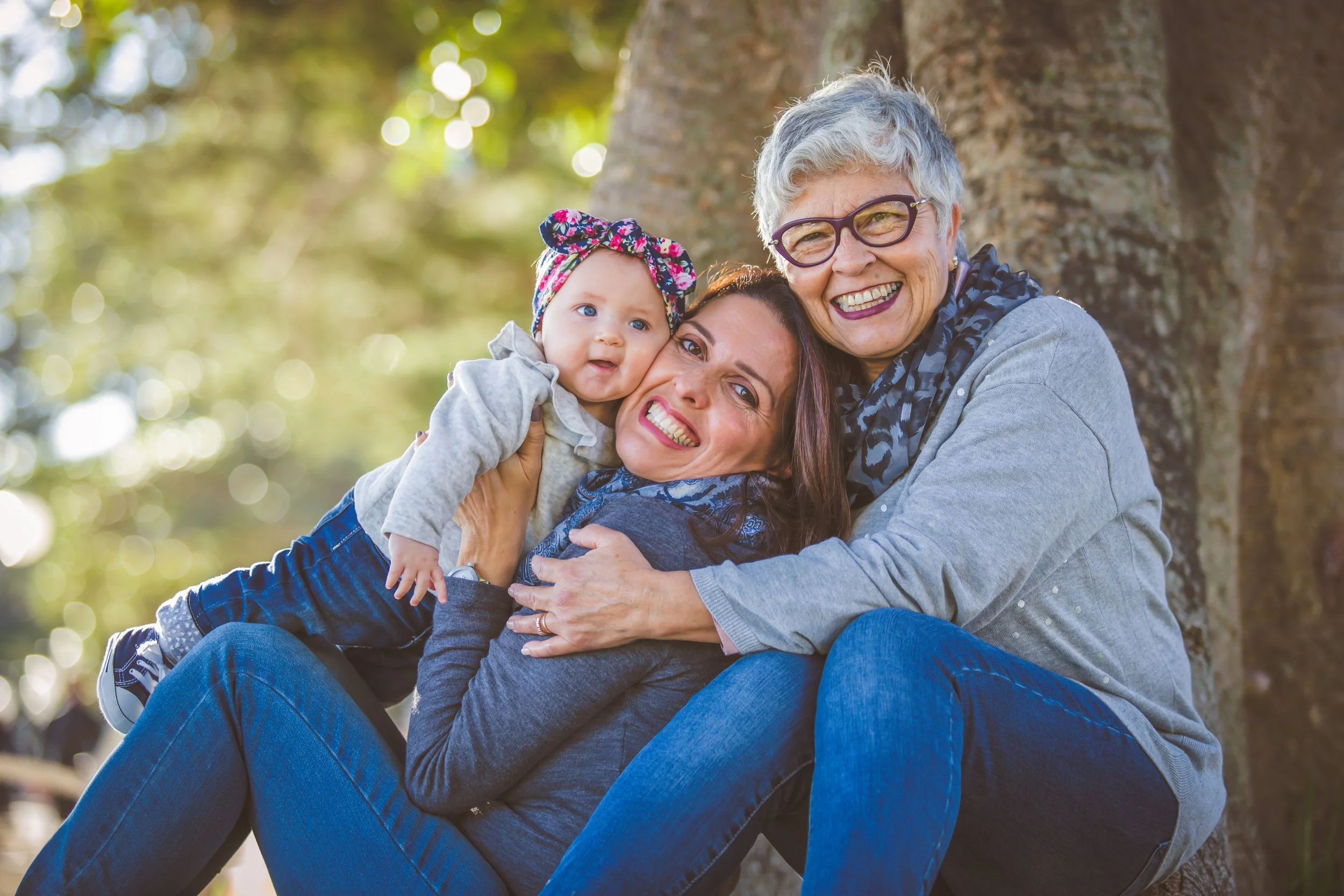 Three generations of women smiling and hugging outdoors near a tree, with sunlight filtering through the leaves.