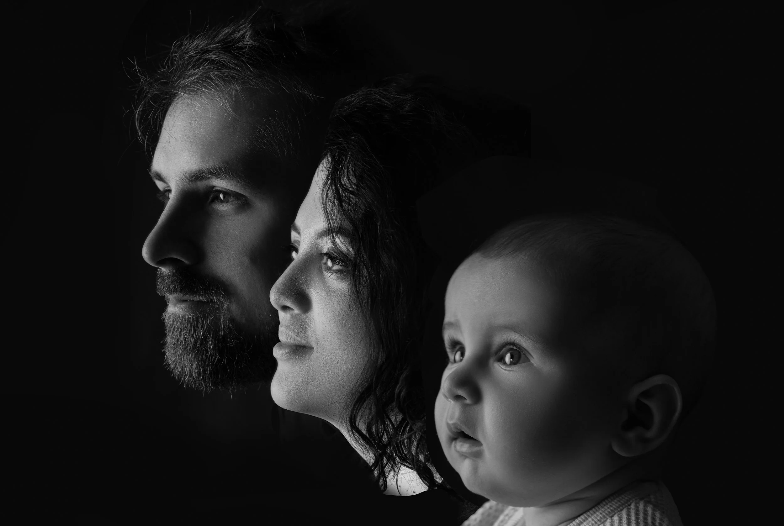 Black and white portrait of a family of three looking to the right, with the man on the left, the woman in the middle, and a young child on the right, all facing the same direction.