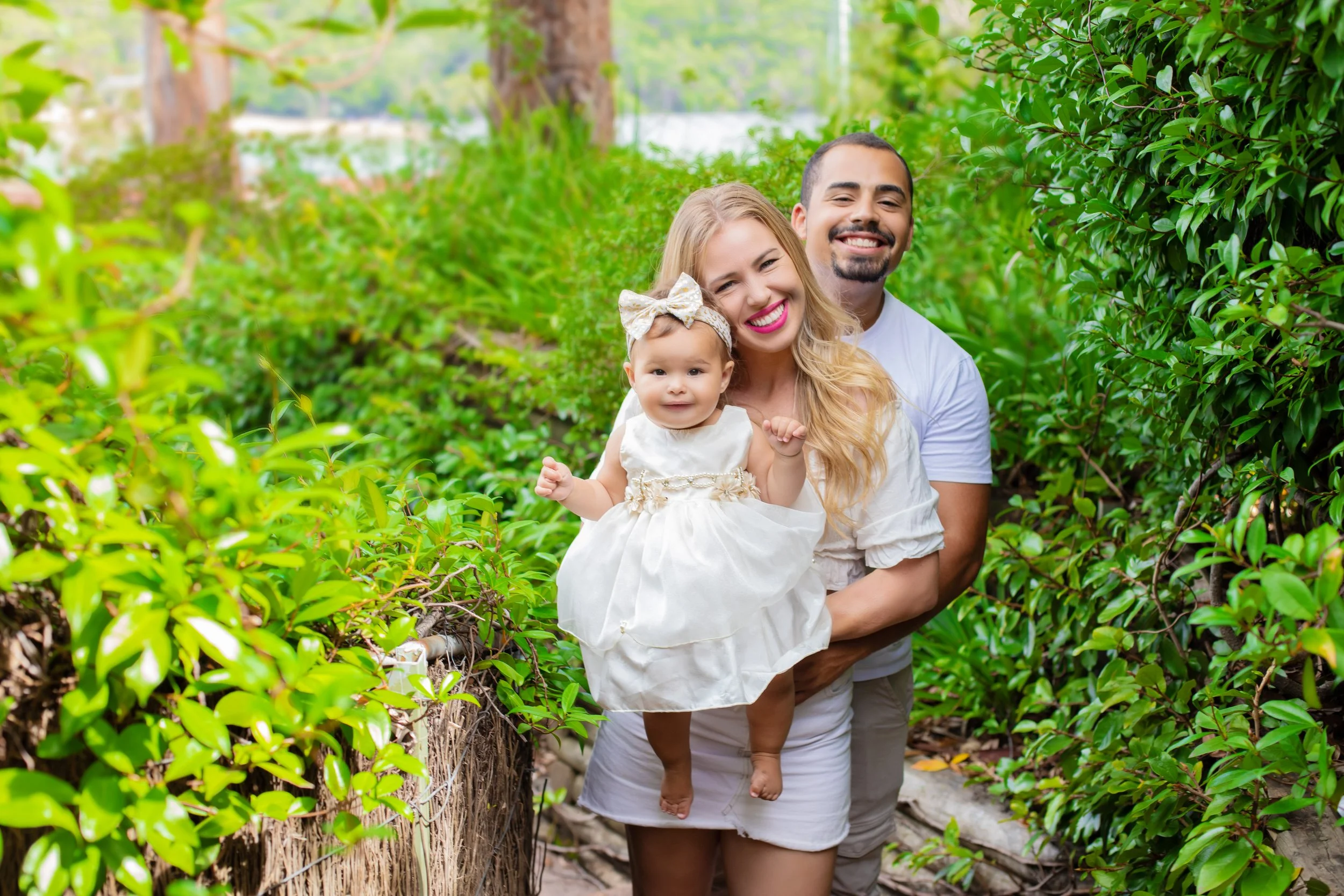 Happy family of three, a woman, a man, and a young girl, smiling and posing outdoors among green bushes and trees.