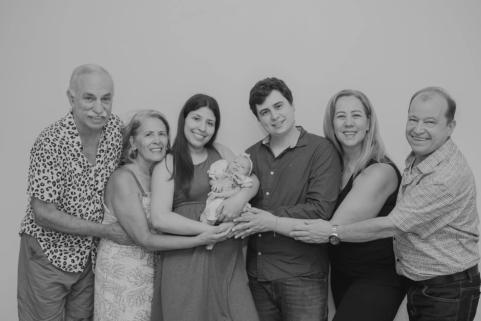 Black and white family photo of seven people, including a newborn baby, smiling and holding the baby, standing against a plain background.