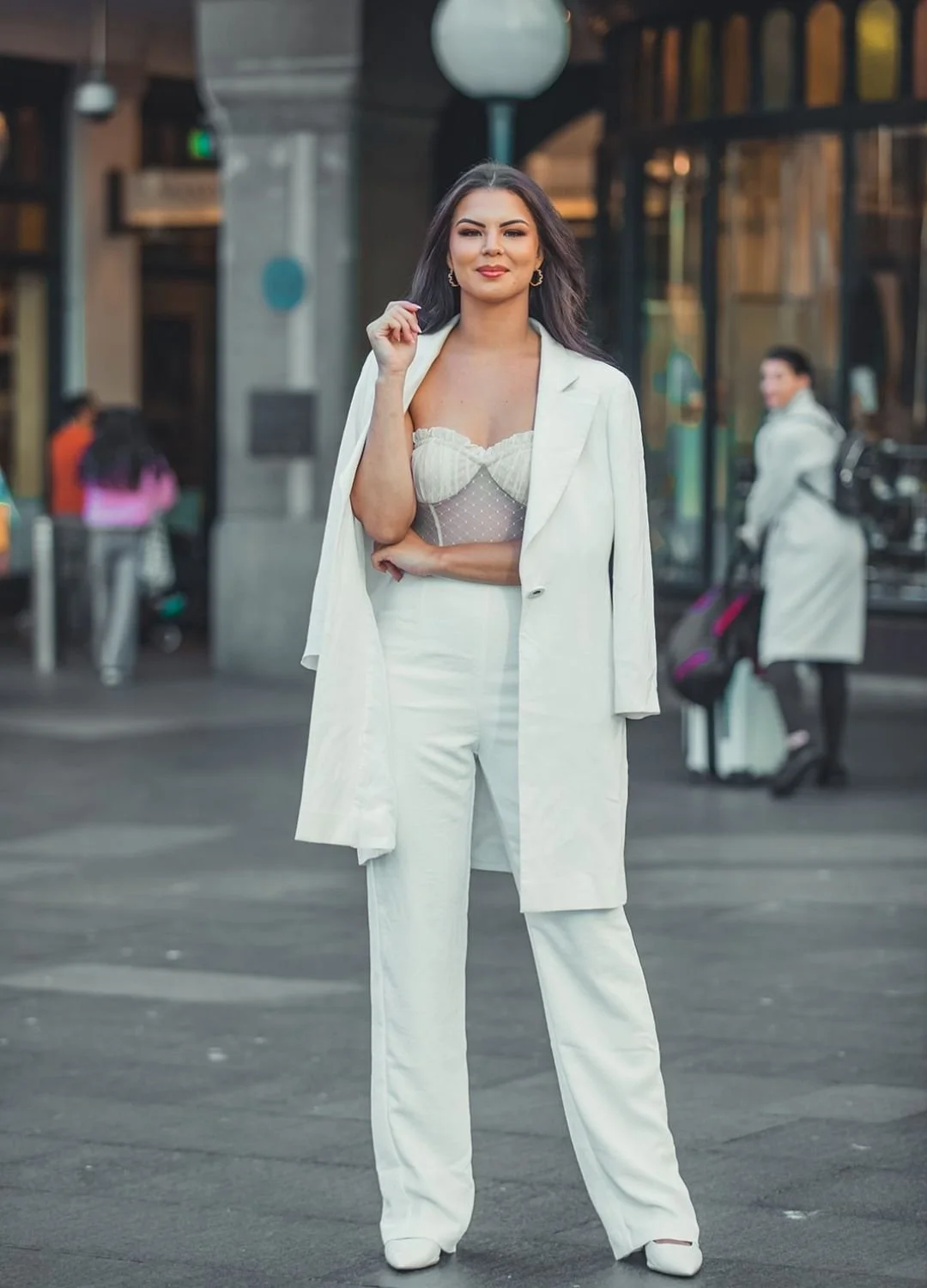 A woman stands confidently on a city street in a white pantsuit with a blazer draped over her shoulders, with a sheer, polka-dotted top underneath.