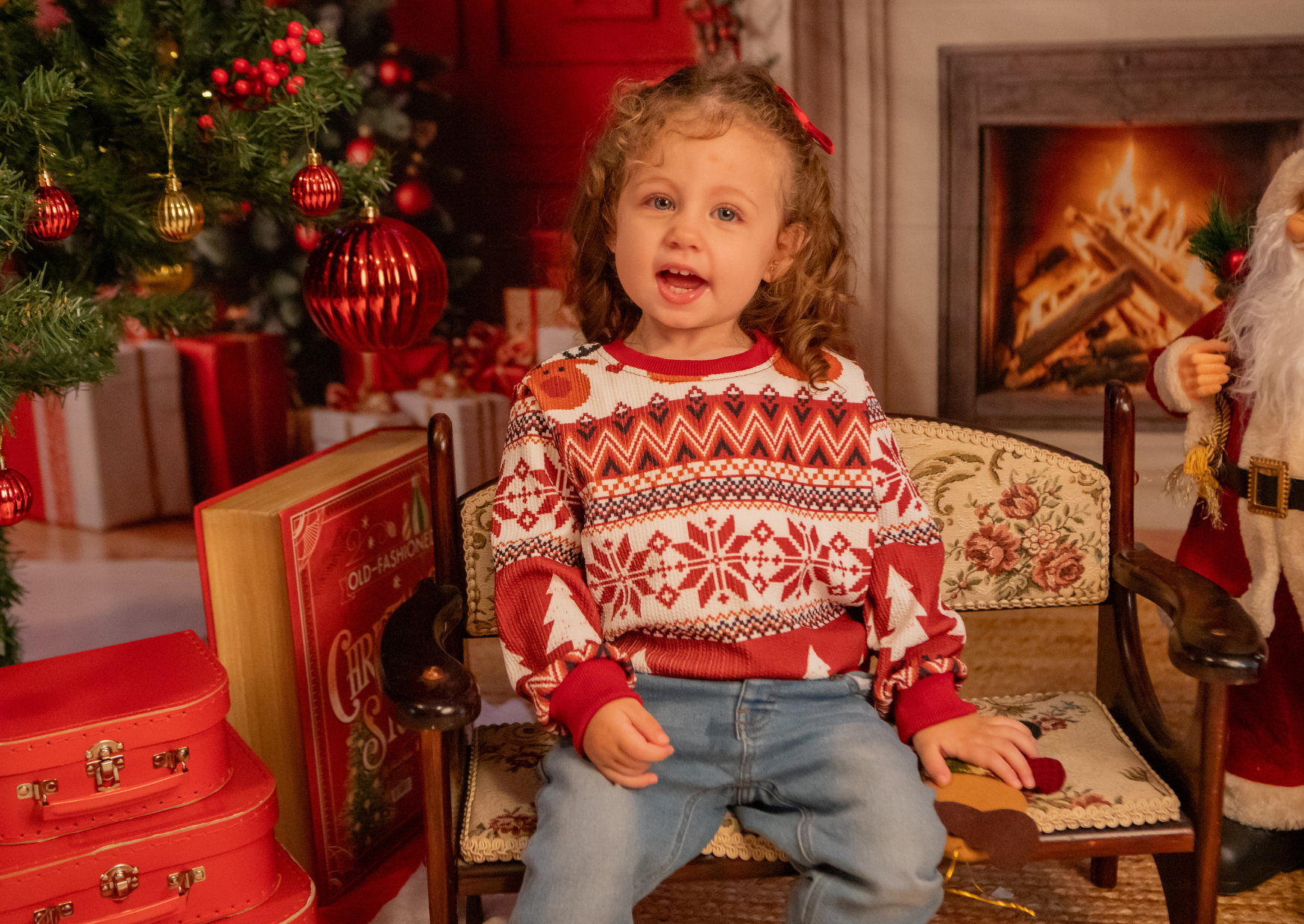 A young girl sitting on a vintage wooden bench with floral upholstery, wearing a red and white Christmas sweater with snowflakes and trees, in a room decorated for Christmas with a lit fireplace, Christmas tree with ornaments, and presents in the bac