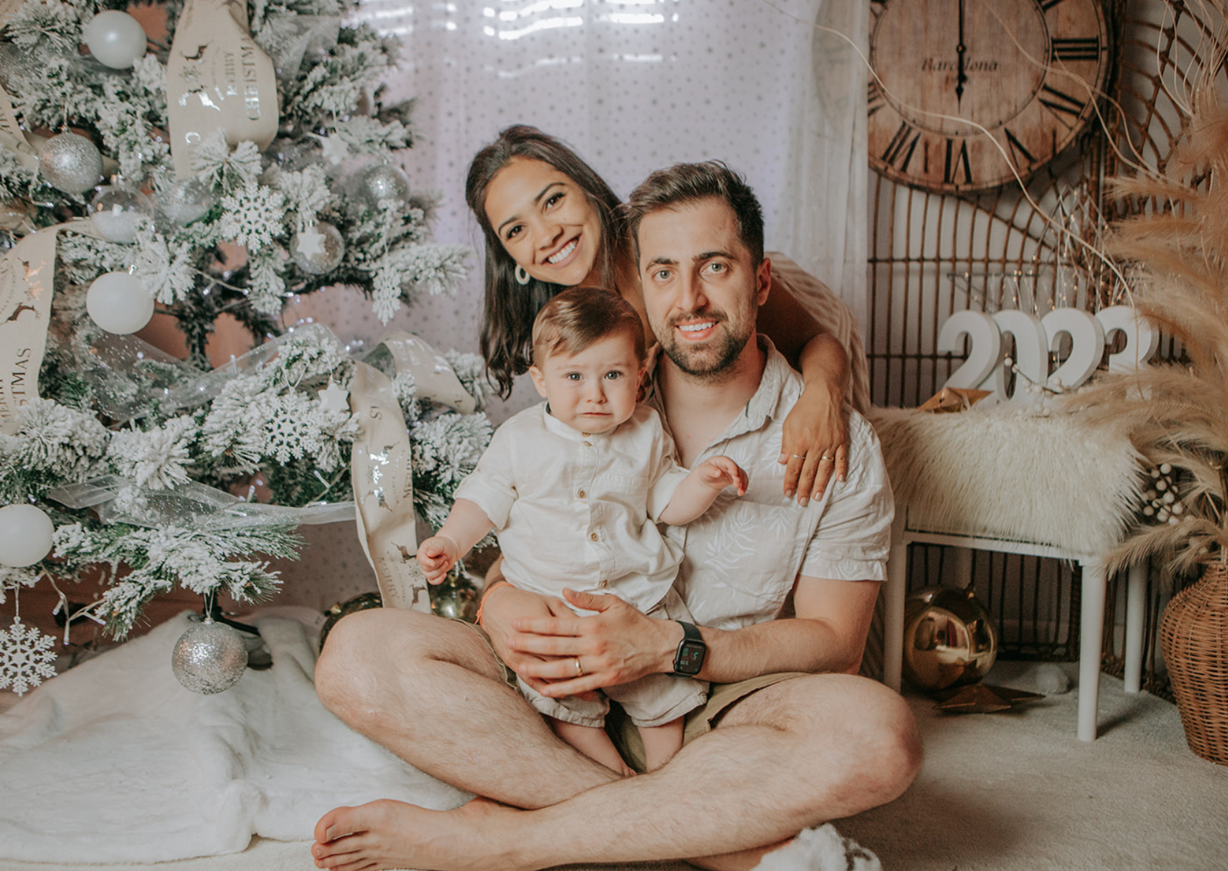 A family of three, including a woman with dark hair, a man with light brown hair, and a young child, sitting on the floor near a decorated Christmas tree. The woman is leaning over from behind, smiling. The man is sitting cross-legged, holding the ch