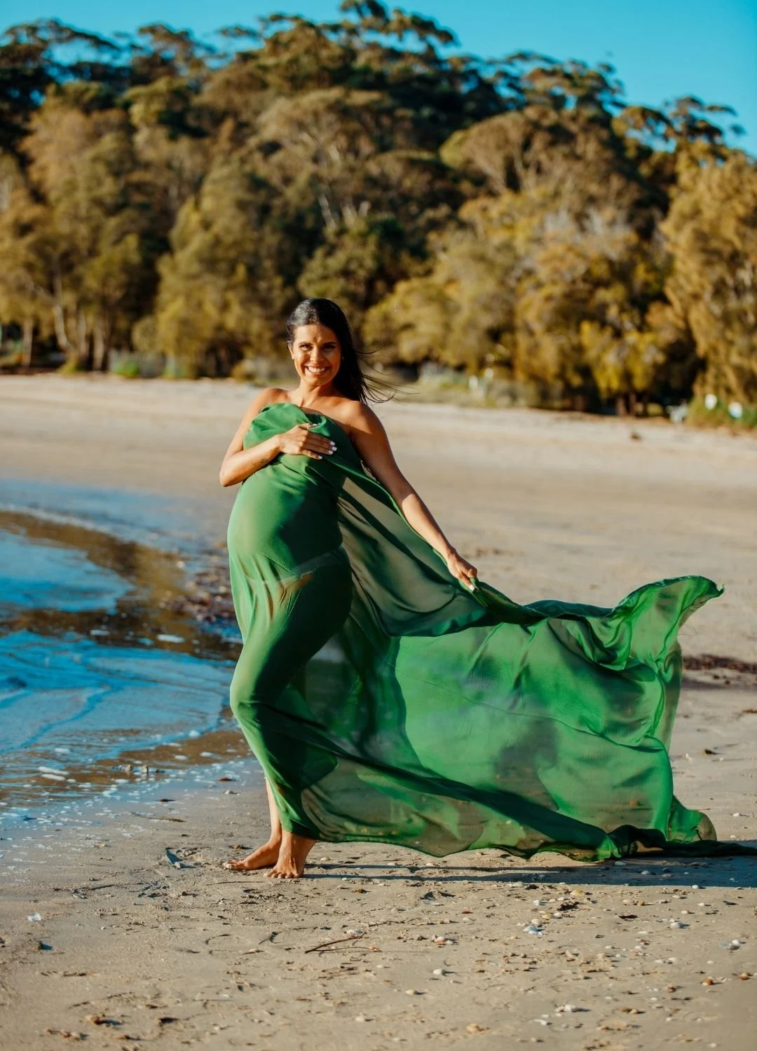 A woman standing barefoot on a sandy beach, wrapped in a flowing green dress, with trees and blue sky in the background.