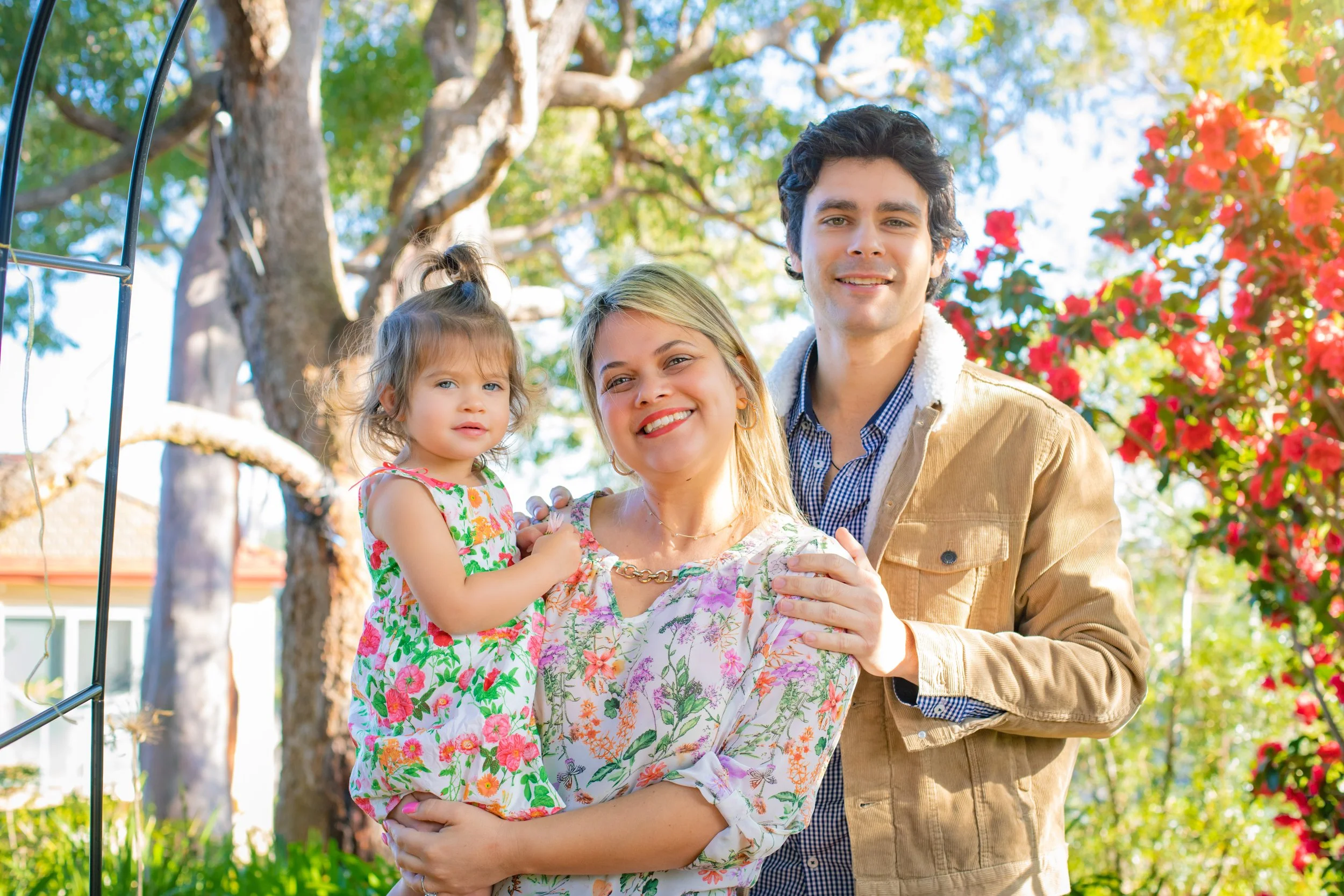 A happy family of three, a woman holding a young girl, standing outdoors in a garden with trees and colorful flowers.