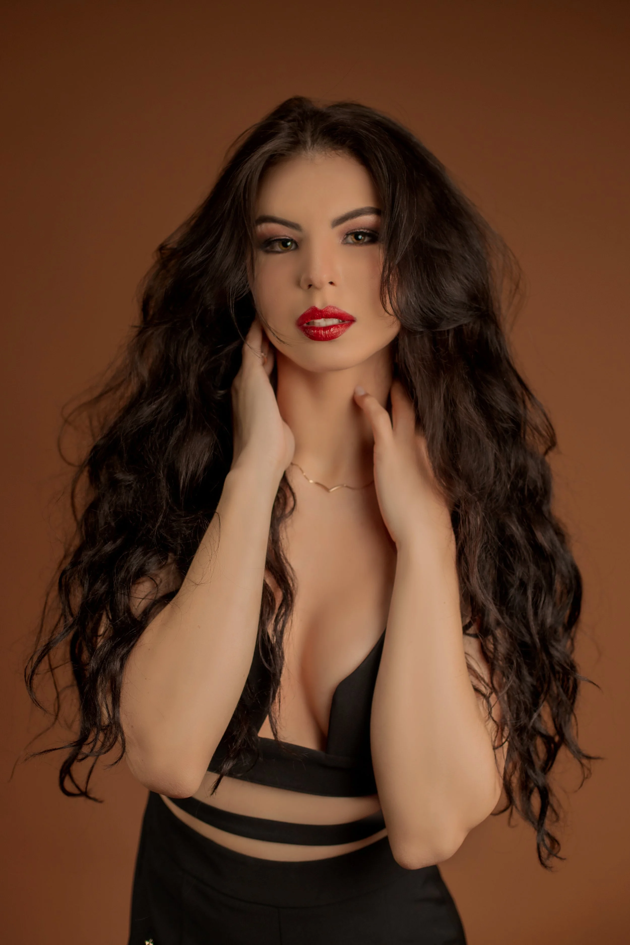 A woman with long, curly dark hair, wearing red lipstick and a black dress with cutouts, poses against a brown background.