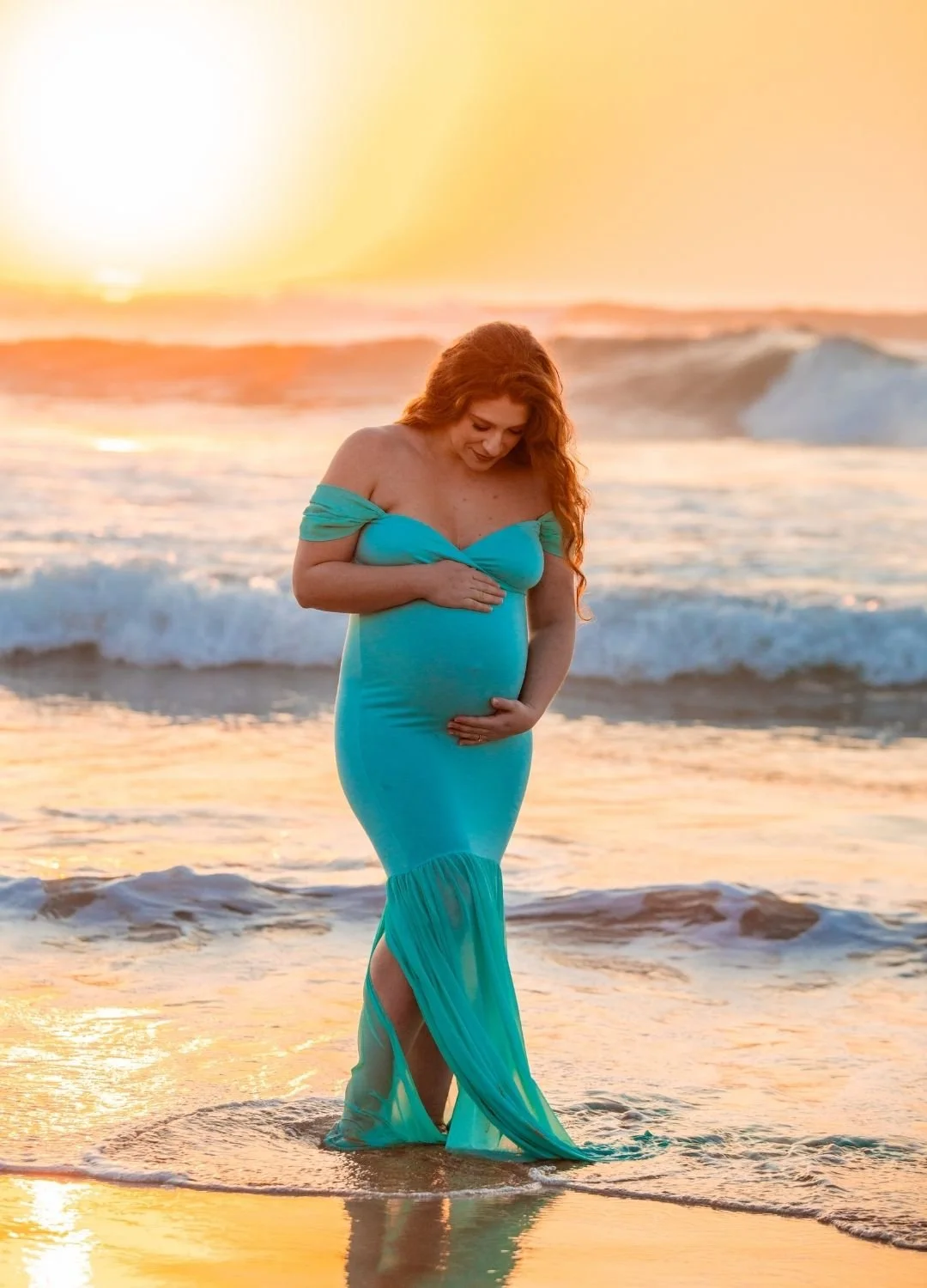 Pregnant woman in a turquoise dress standing in the ocean at sunset.