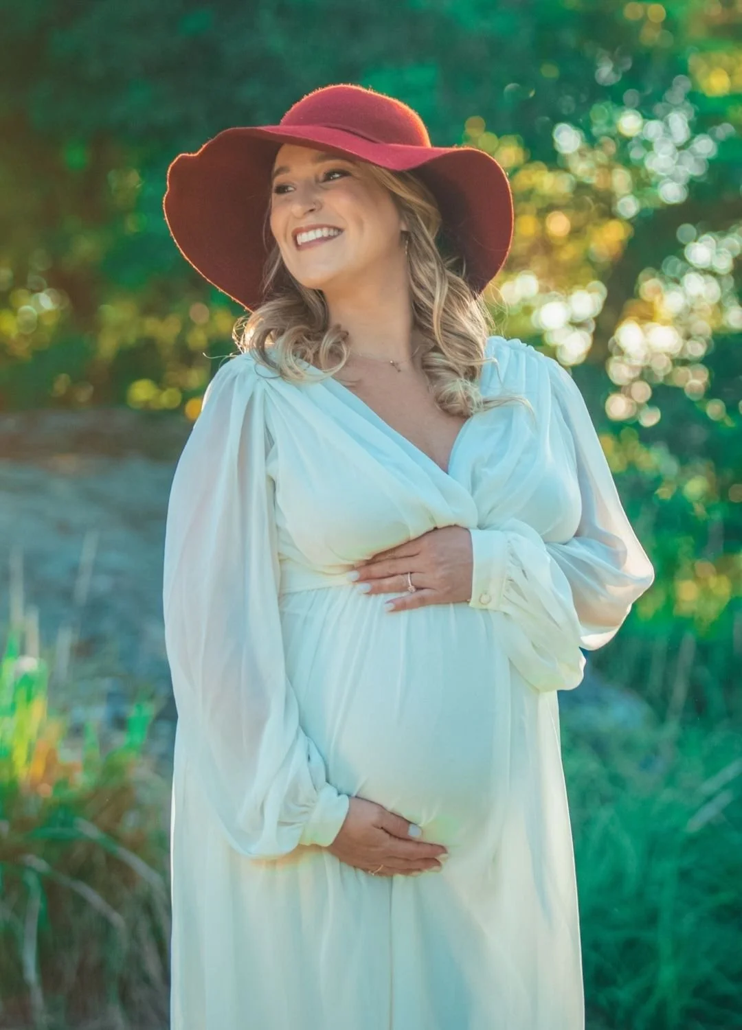 A pregnant woman in a white dress and a large red hat, smiling outdoors with green foliage in the background, holding her belly.