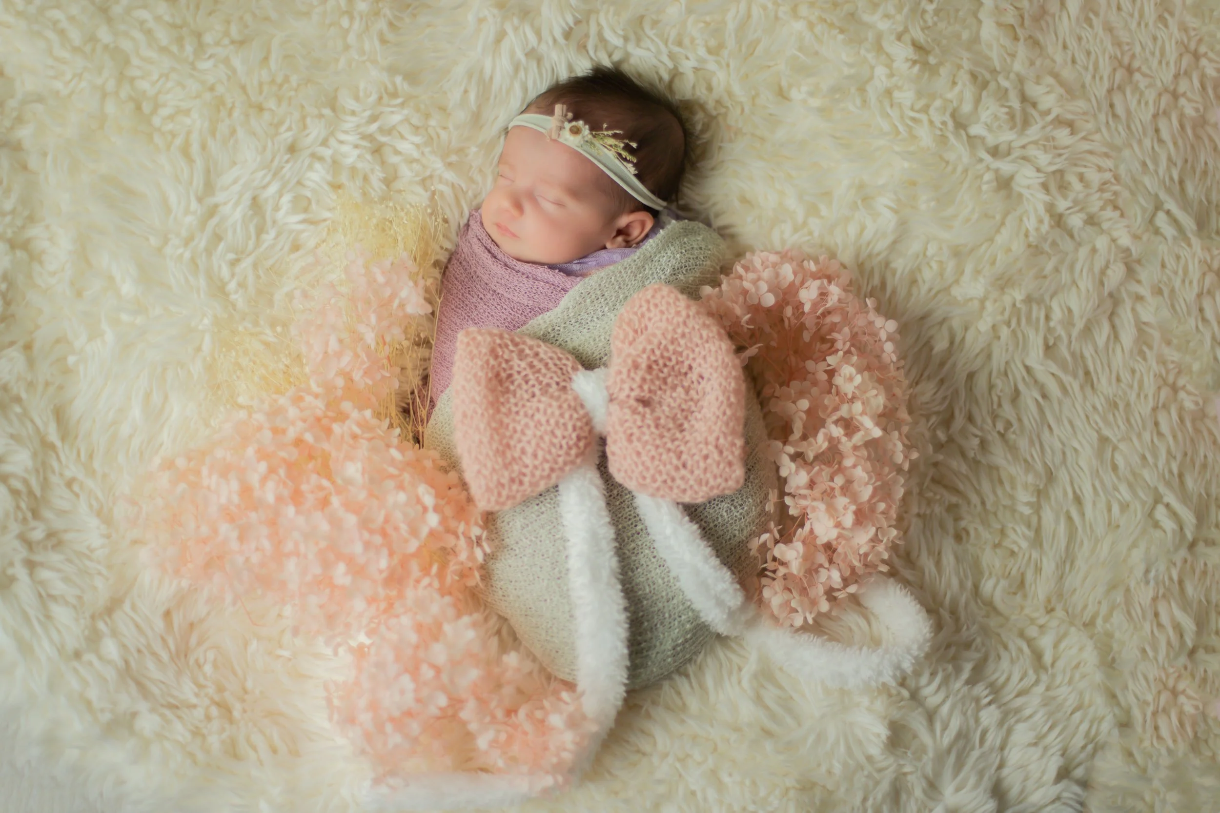 A sleeping newborn baby swaddled in a pink and gray blanket, wearing a small flower headband, lying on a soft cream fuzzy surface surrounded by pink and peach artificial flowers and a large pink knitted bow.
