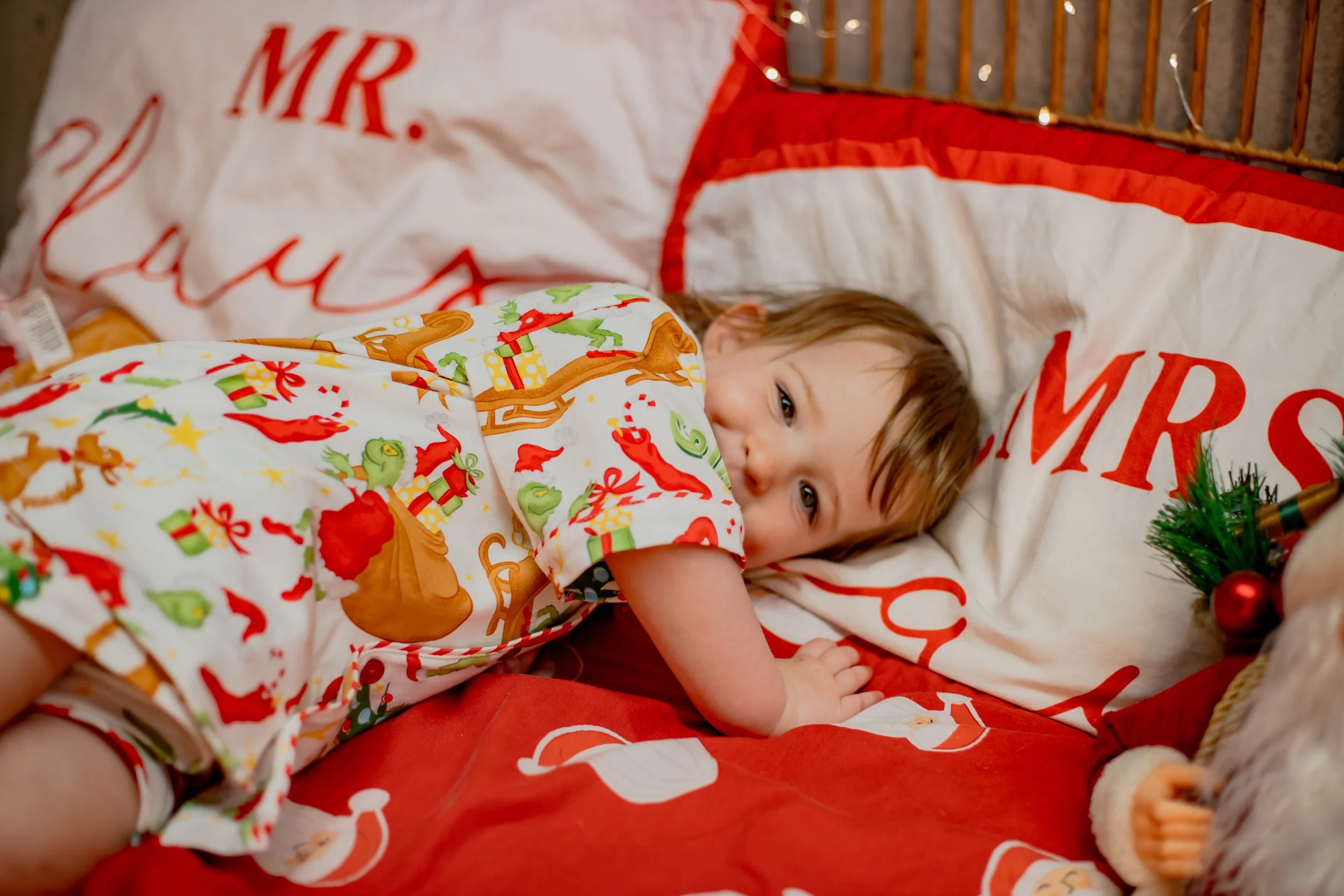 A young child lying on a bed with Christmas-themed bedding, partially hiding face behind a pillow that says 'MERRY CHRISTMAS', wearing holiday pajamas with reindeer and Christmas tree patterns, near Christmas decorations.