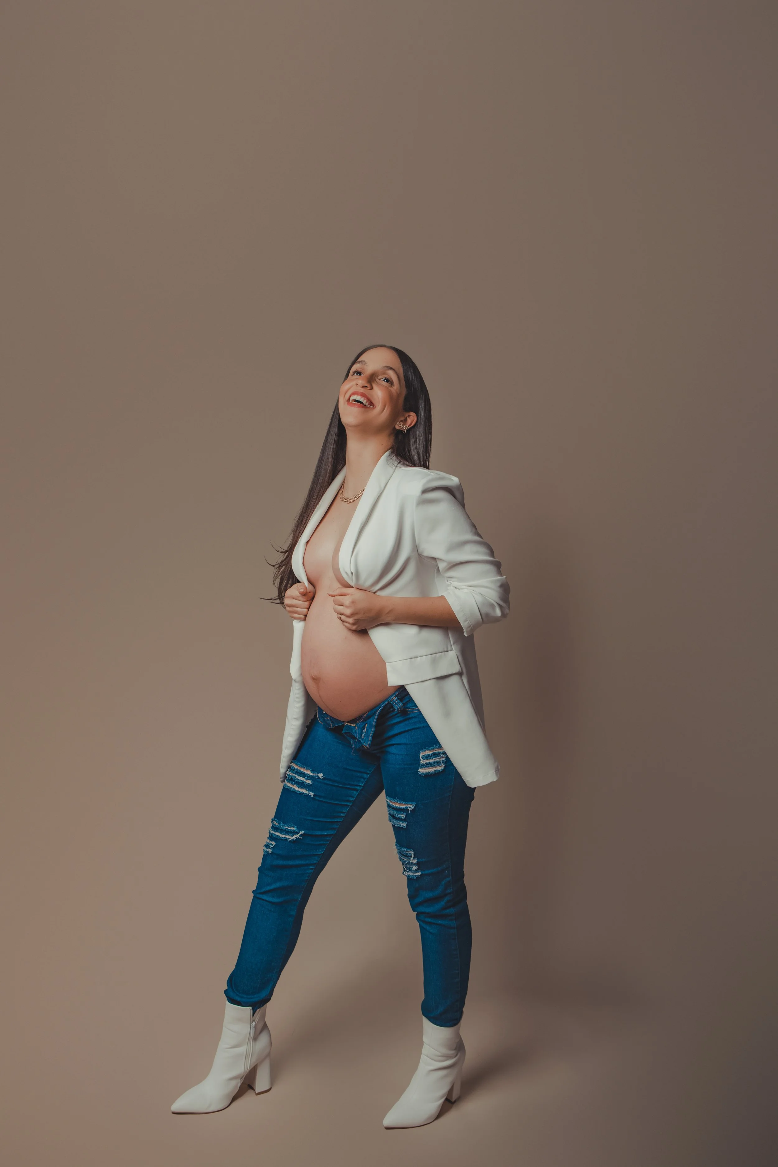 A woman with long dark hair, wearing a white blazer, distressed blue jeans, and white high-heeled boots, stands against a neutral background, smiling with her hands on her open blazer to reveal her pregnant belly.