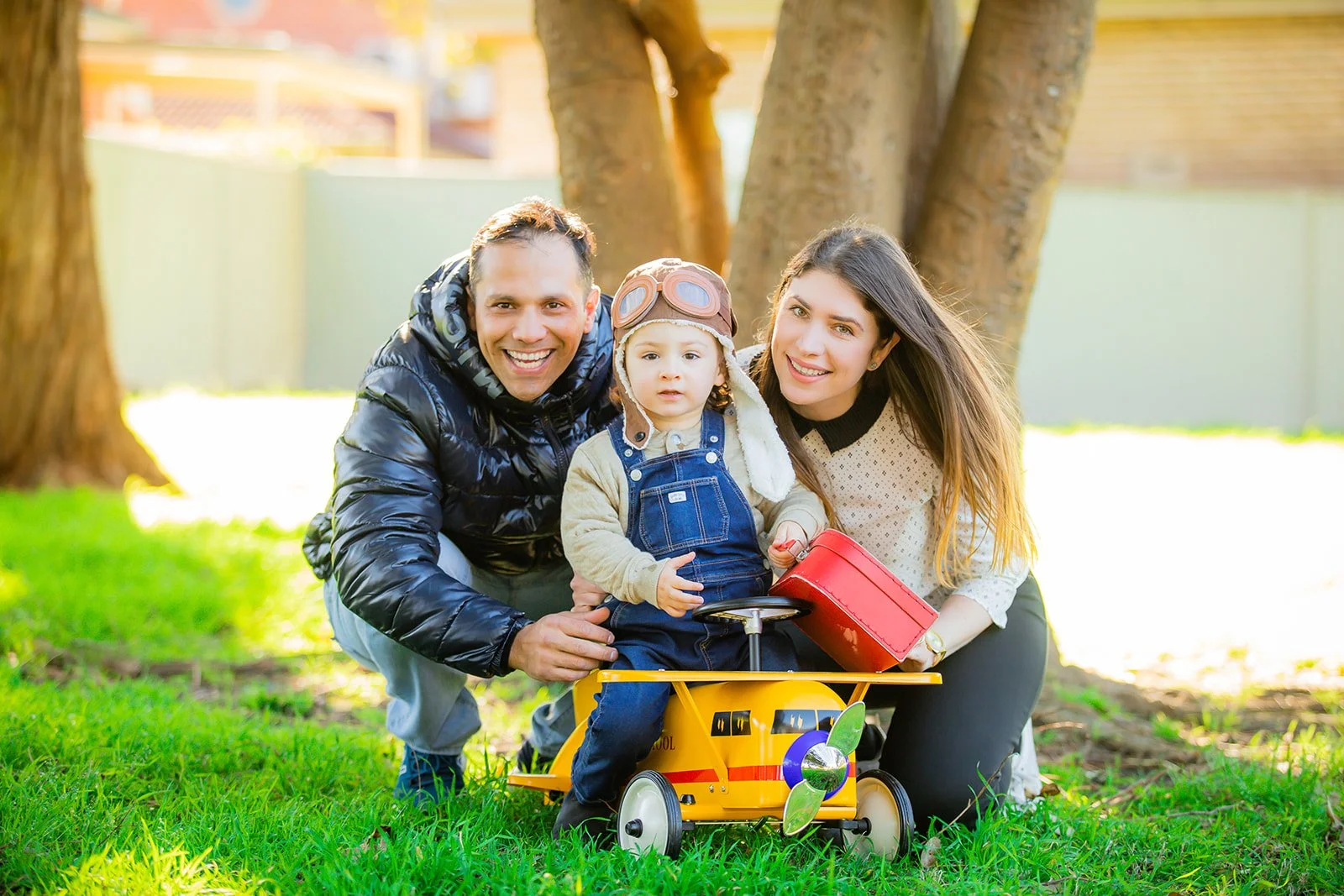 A happy family of three: a father, mother, and young child, smiling and sitting on the grass outdoors. The child is sitting in a yellow pedal airplane toy, wearing aviator goggles and a helmet. The family is surrounded by trees in a park setting.