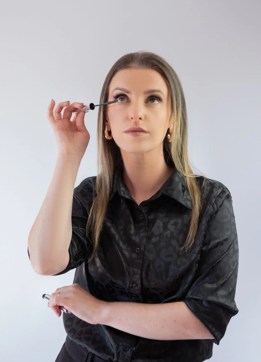 A woman with long brown hair, wearing a black patterned shirt, applying mascara to her eyelashes with a mascara wand while looking slightly upward.