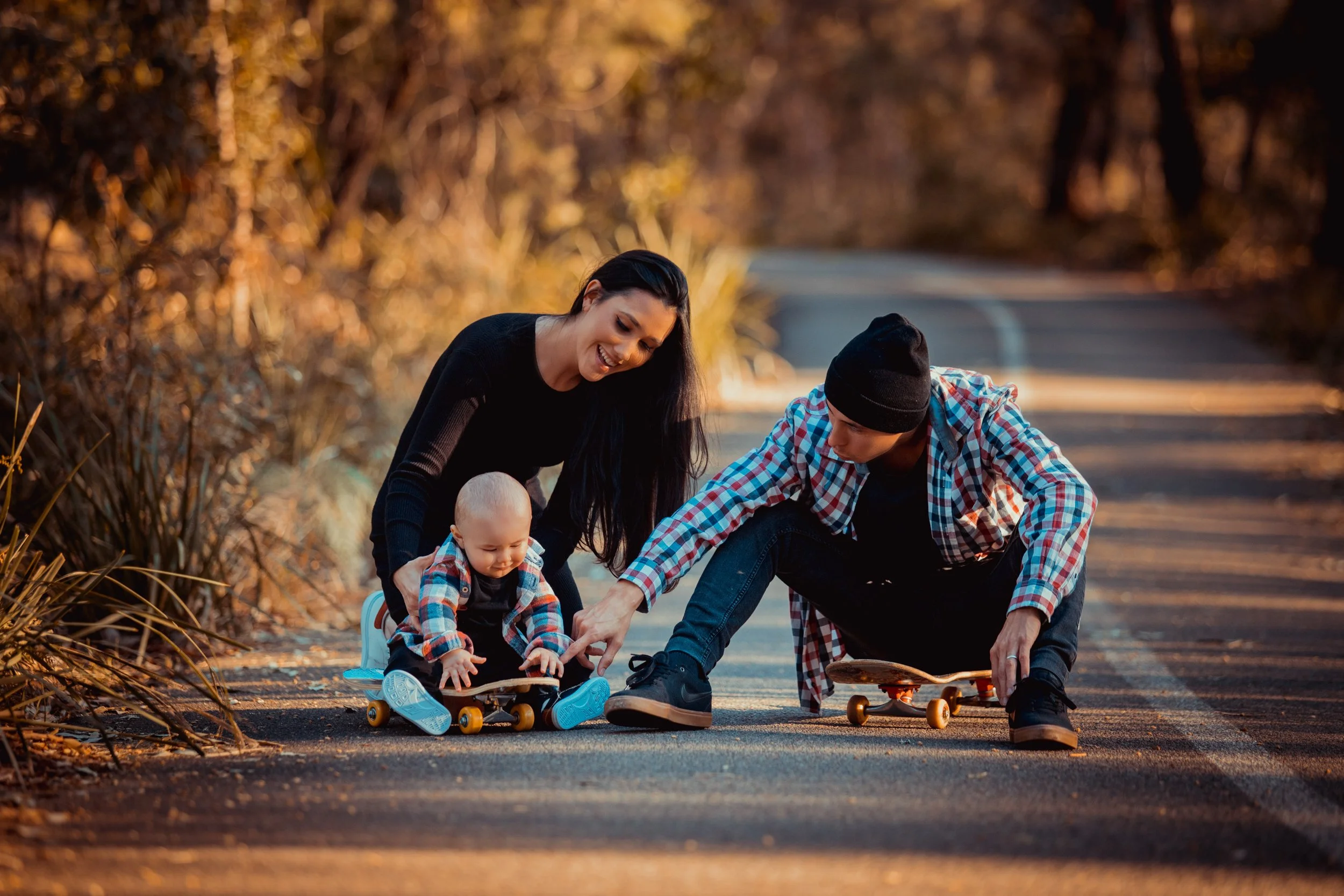 Family of three, including a woman and a man, outdoors on a deserted road during autumn, helping a young child on a skateboard, all smiling and enjoying the moment.