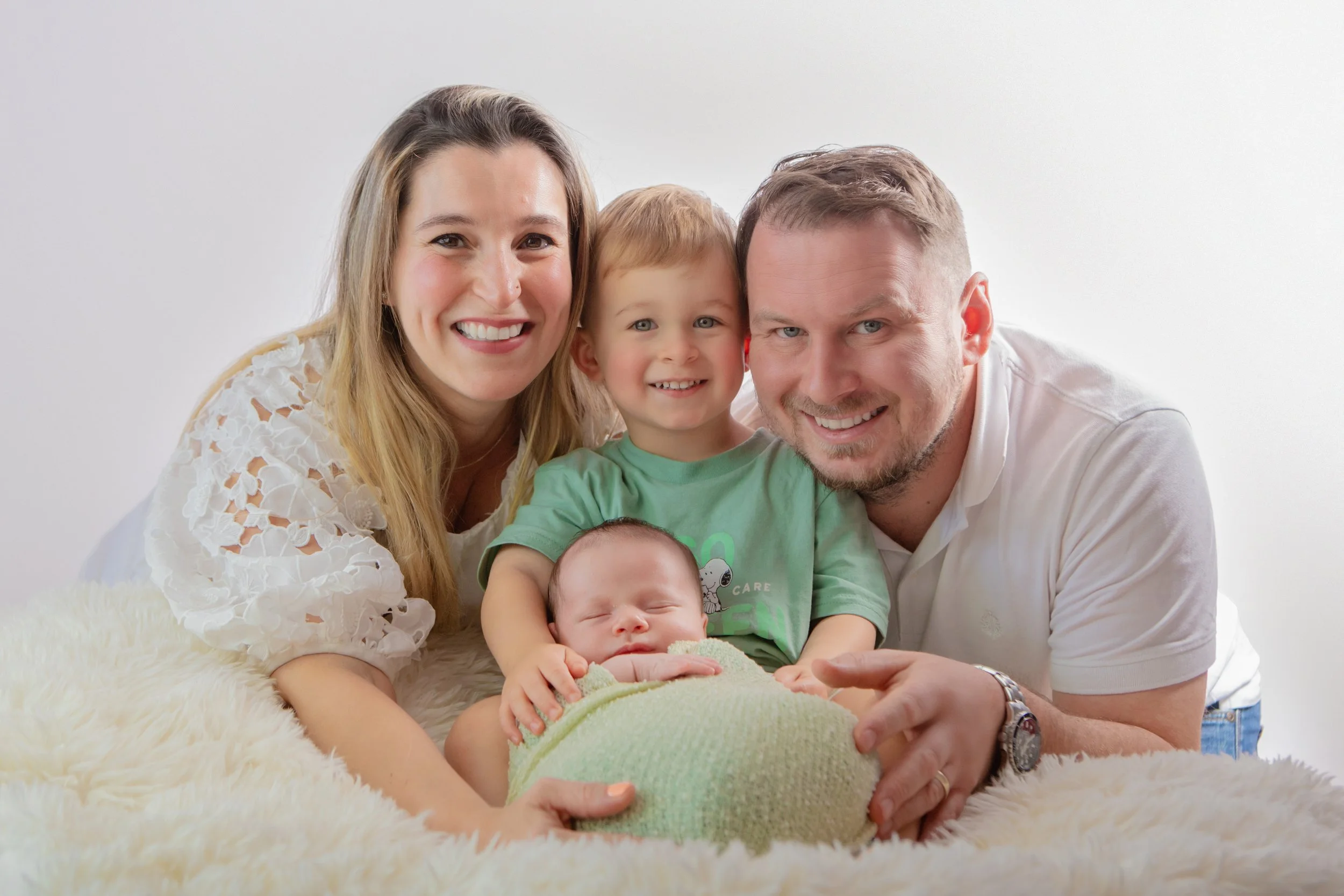 Family of four smiling at camera, lying on a fluffy cream-colored blanket. The mother has long blonde hair and wears a white lace top. The father has short light brown hair and a beard, wearing a white polo shirt. Their young son has short blonde hai