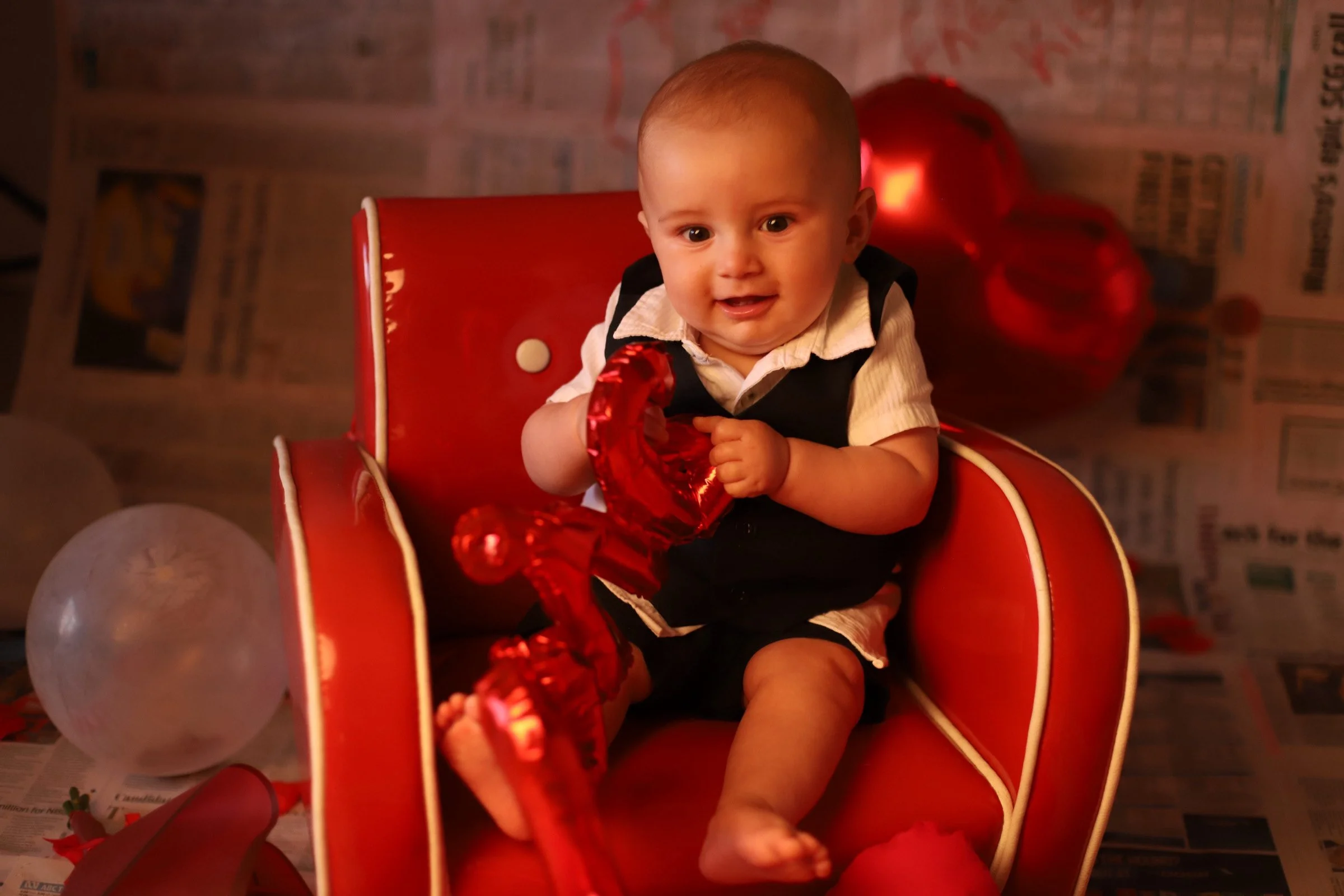 A cute baby sitting on a red chair, holding a red balloon animal, with decorative balloons and balloons on the floor, and newspaper-covered flooring in the background.