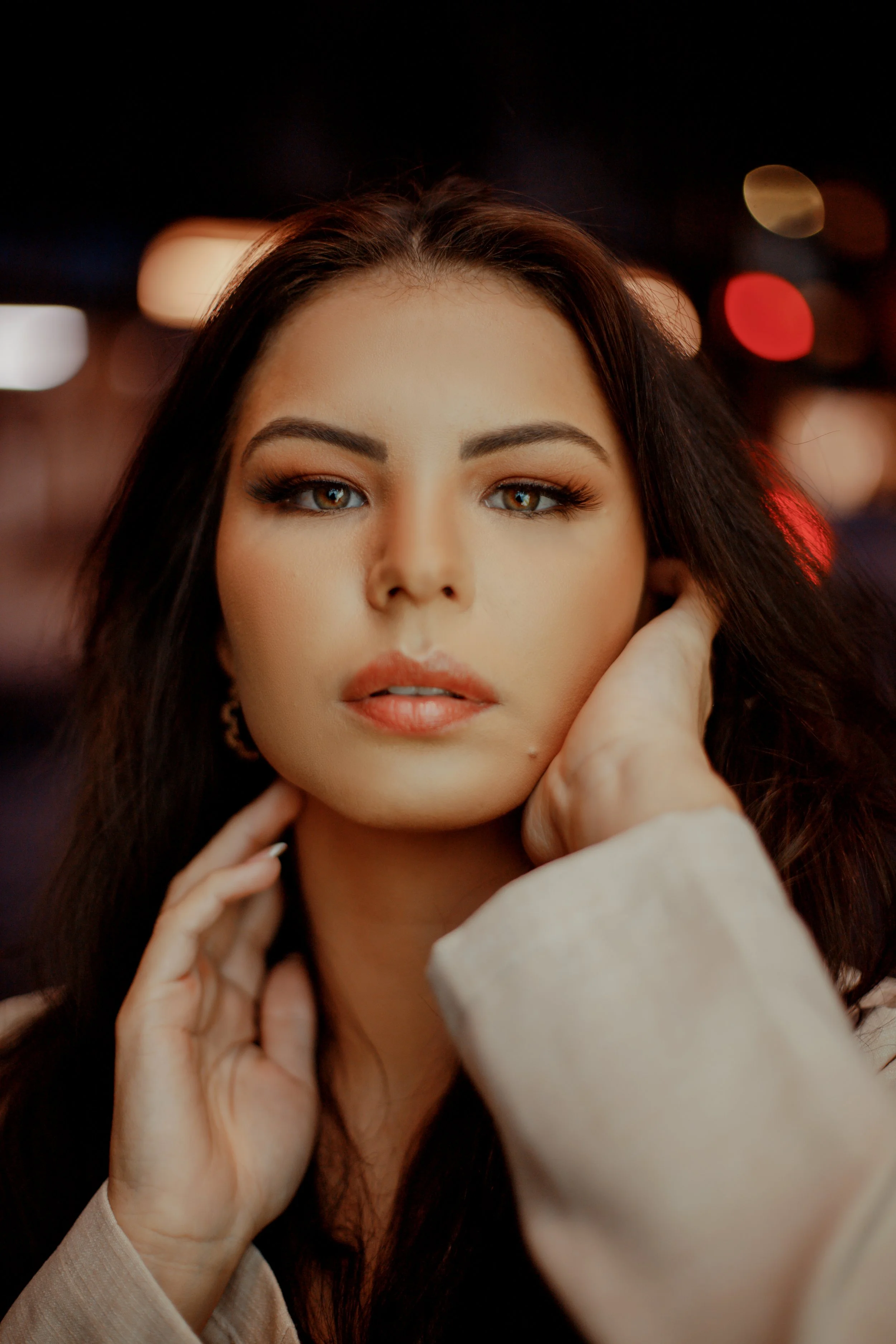 A close-up portrait of a woman with long dark hair, wearing makeup, and gazing directly at the camera with a confident expression, set against a night cityscape with blurred colorful lights.