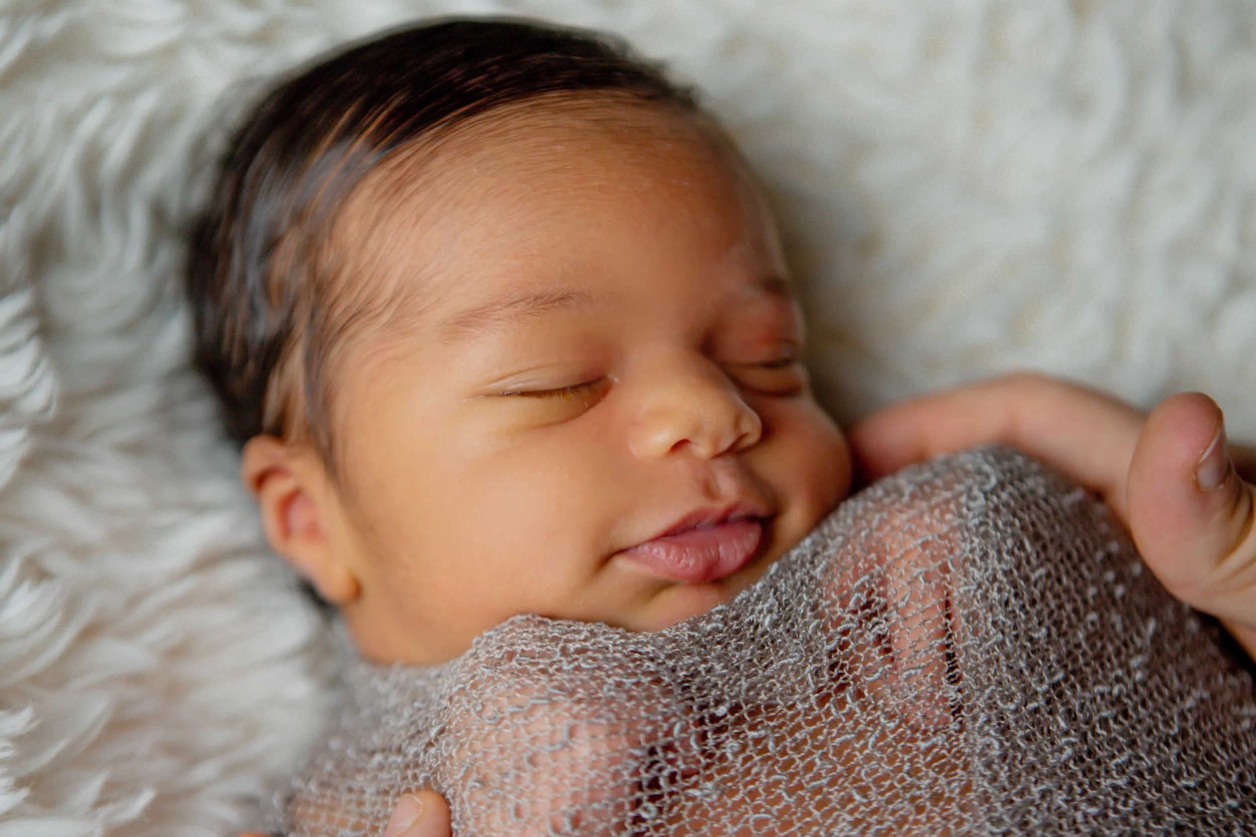 Close-up of a sleeping baby with dark hair, wrapped in a gray netted blanket, resting on a soft, white textured surface.