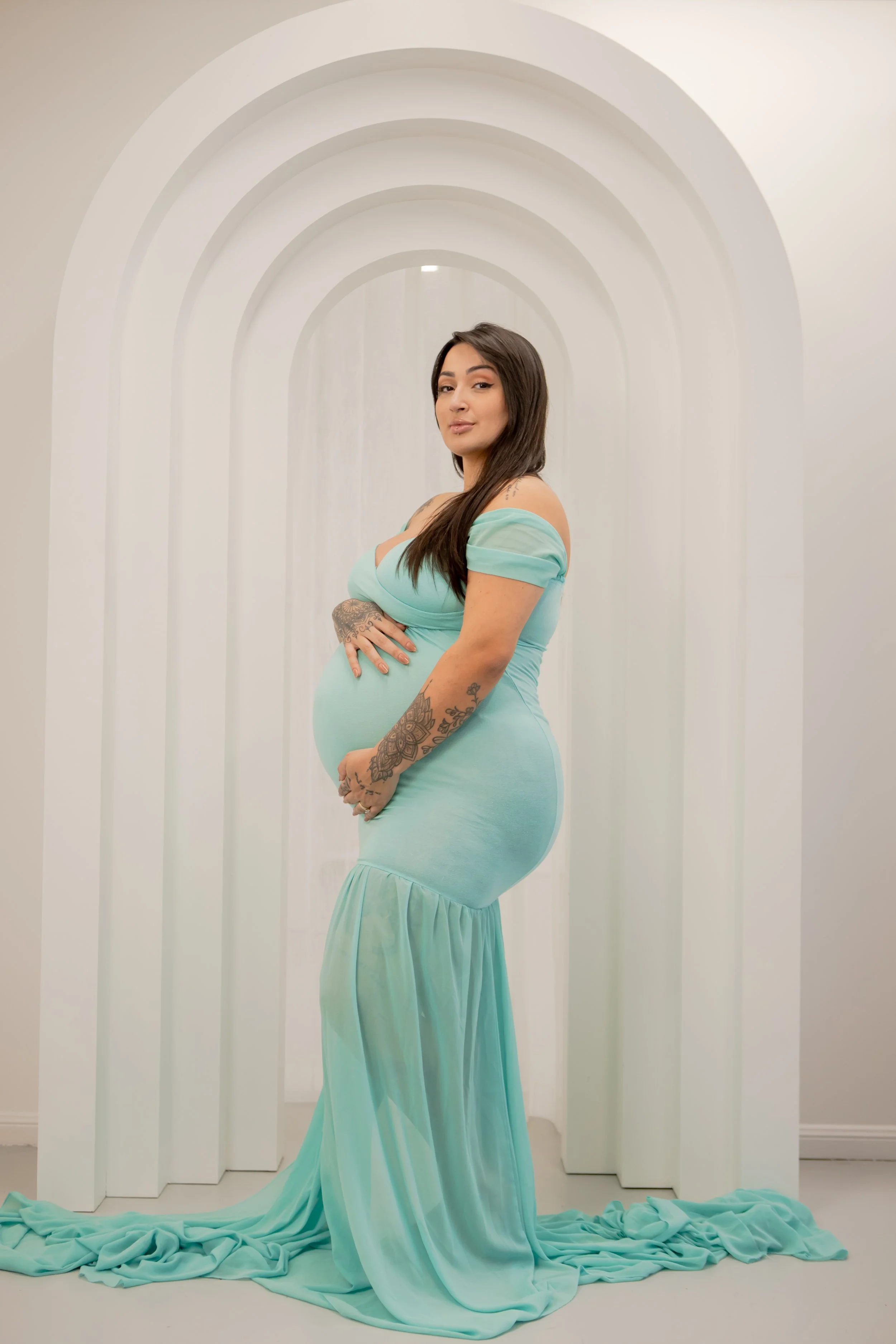 Pregnant woman in a light blue dress standing in front of a white, arched backdrop.