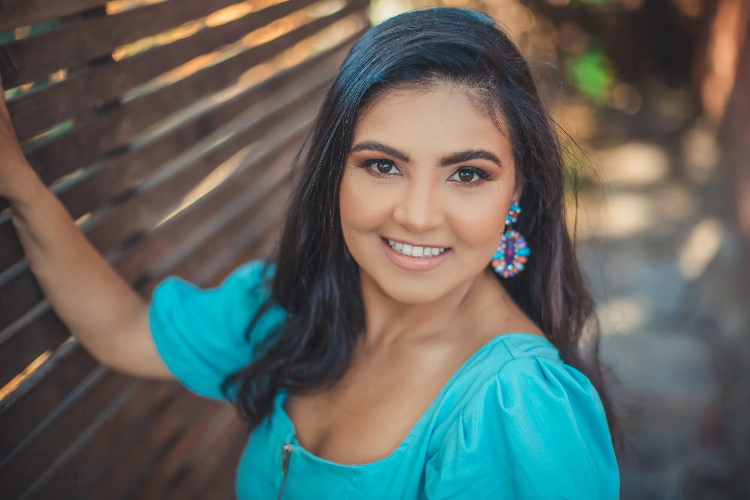 A young woman with dark hair and brown eyes, smiling and wearing a bright blue top with colorful earrings, standing outdoors near a wooden fence.