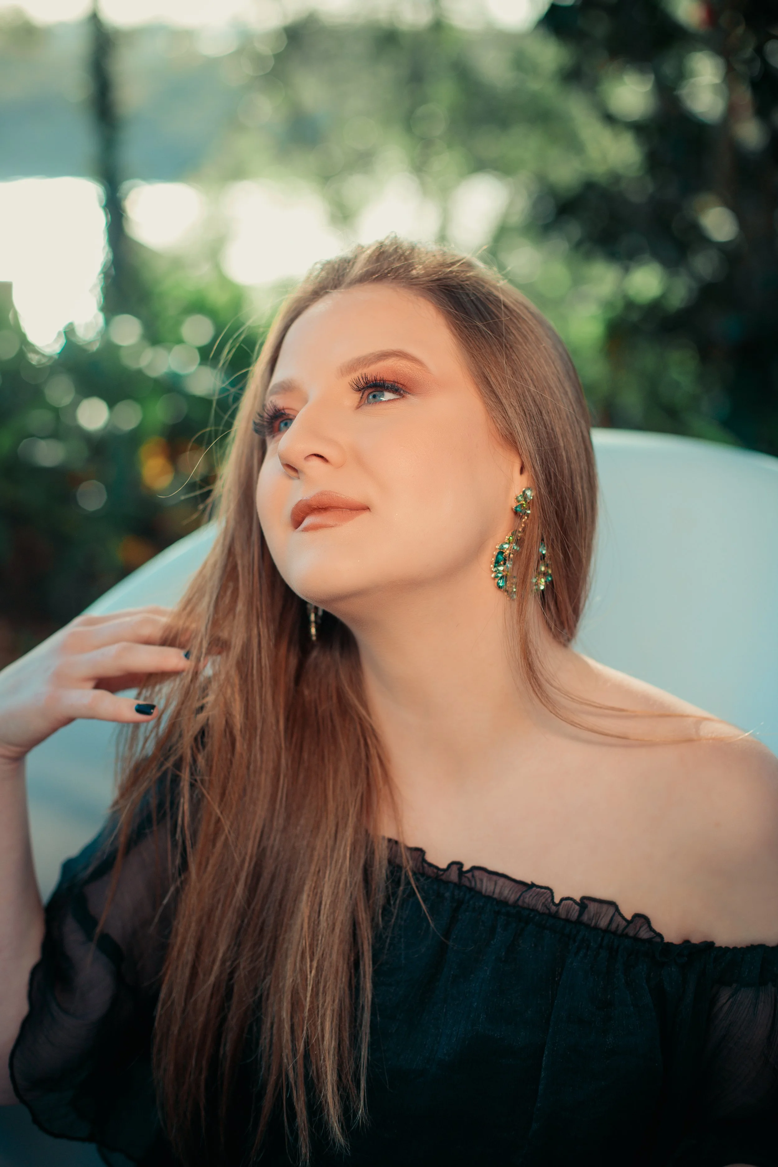 A woman with long brown hair and blue eyes, wearing gold earrings with green stones, rests her hand on her neck and looks slightly upward, outdoors with blurred green foliage in the background.