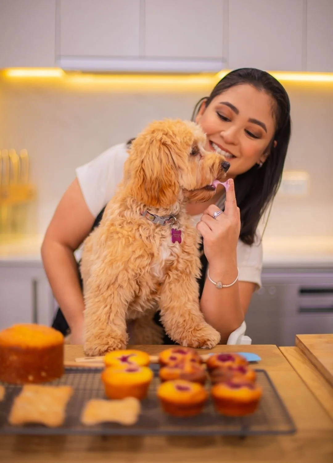 A woman with dark hair and a white shirt happily touching her dog's nose in a kitchen, with cupcakes and baked goods on the table in front of them.