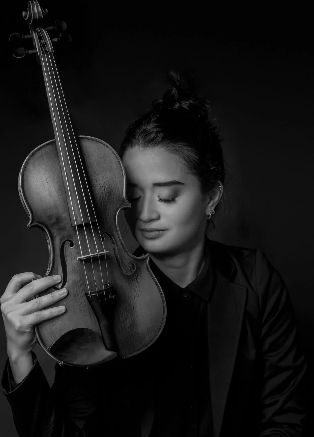 A woman with dark hair styled in a top knot holding a violin against her face with eyes closed, in black and white photography.