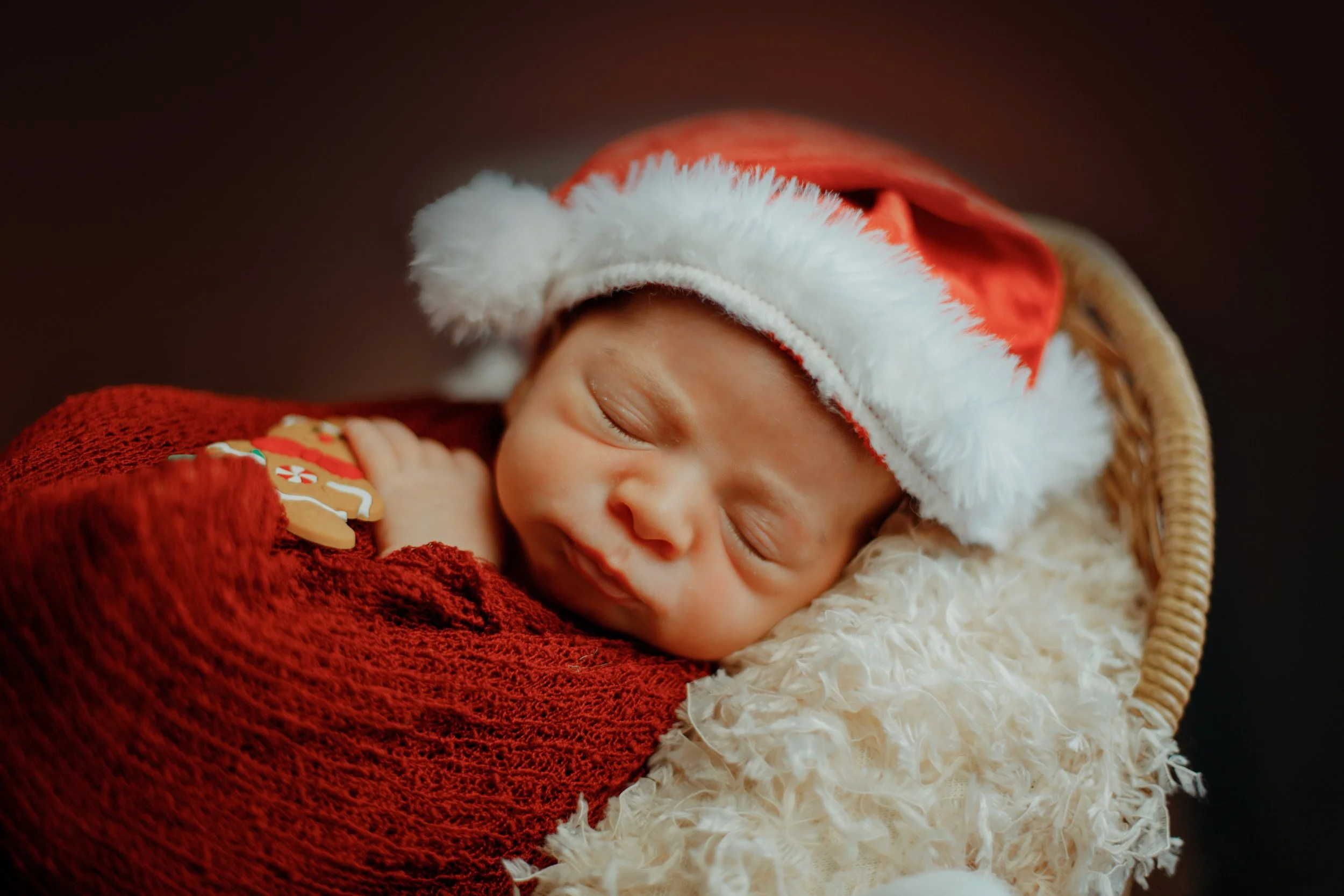 A newborn baby sleeping, wearing a Santa hat and a red blanket, lying on a fluffy cream-colored blanket.