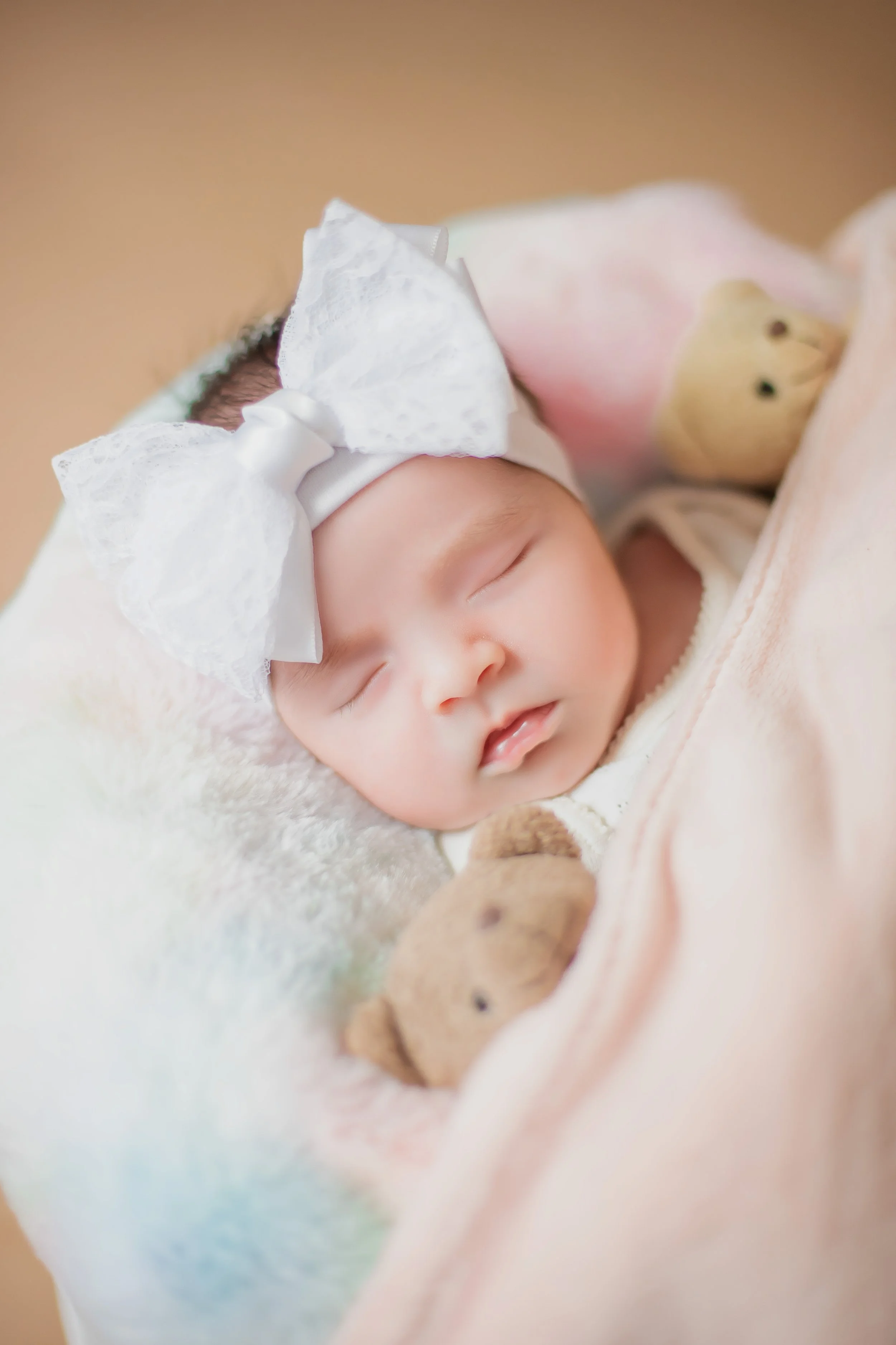 A sleeping baby girl with a large white bow headband, wrapped in a soft pink blanket, and holding a small teddy bear.