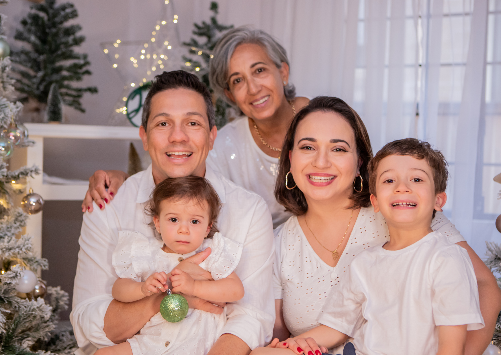 Family celebrating Christmas indoors, with decorated Christmas trees and string lights in the background.