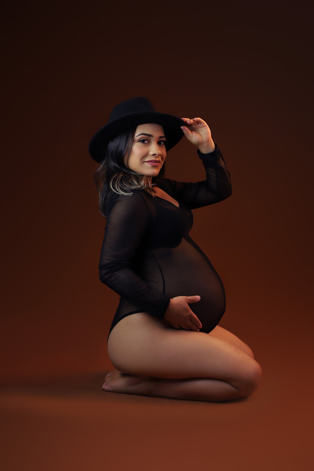 A pregnant woman kneeling on the floor, wearing a black sheer outfit and a black hat, smiling and touching her belly against a dark brown background.