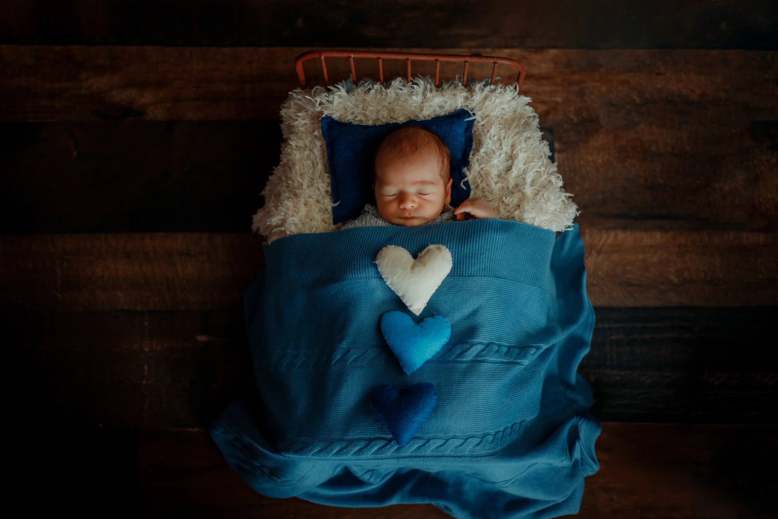 A sleeping baby lying in a small bed with a dark wooden floor background, decorated with plush hearts in white, light blue, and dark blue.