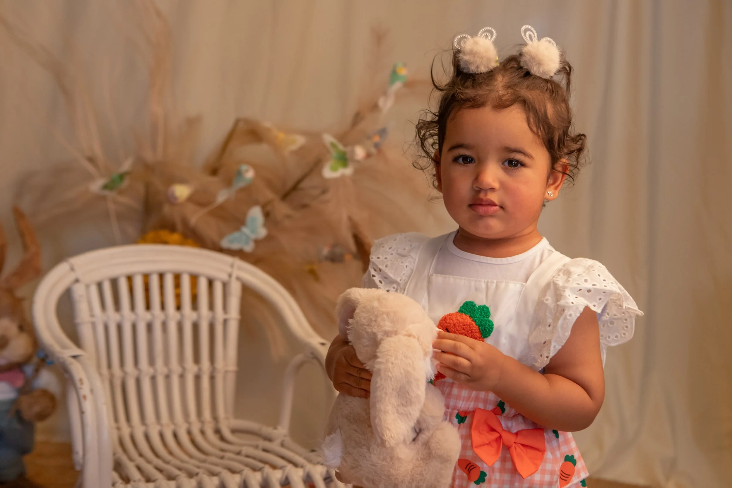 A young girl with curly hair and a white dress holding a stuffed animal, standing indoors with decorative feathers and butterfly ornaments in the background.