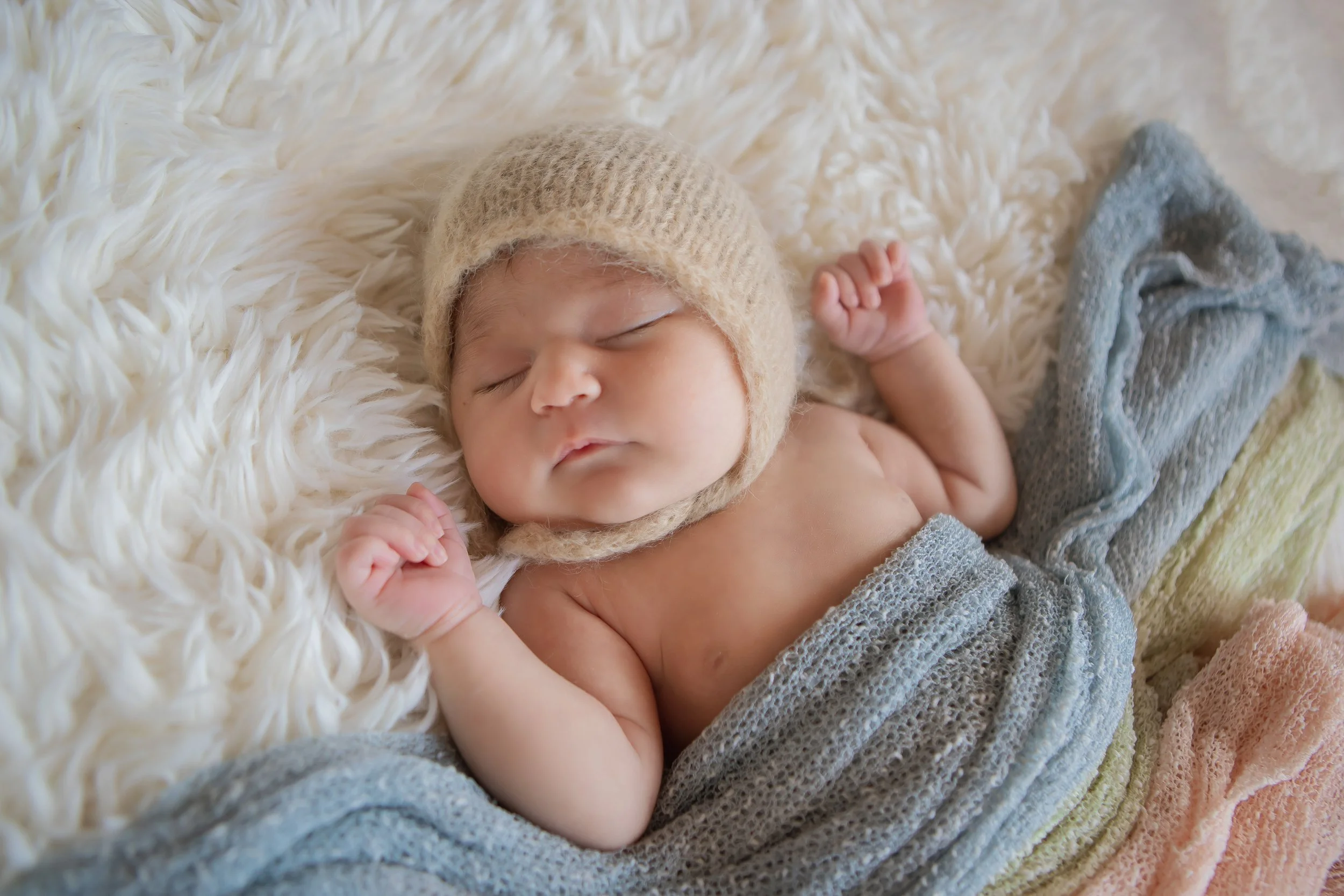 Sleeping baby wearing a knit hat and wrapped in a colorful blanket on a fluffy white surface.