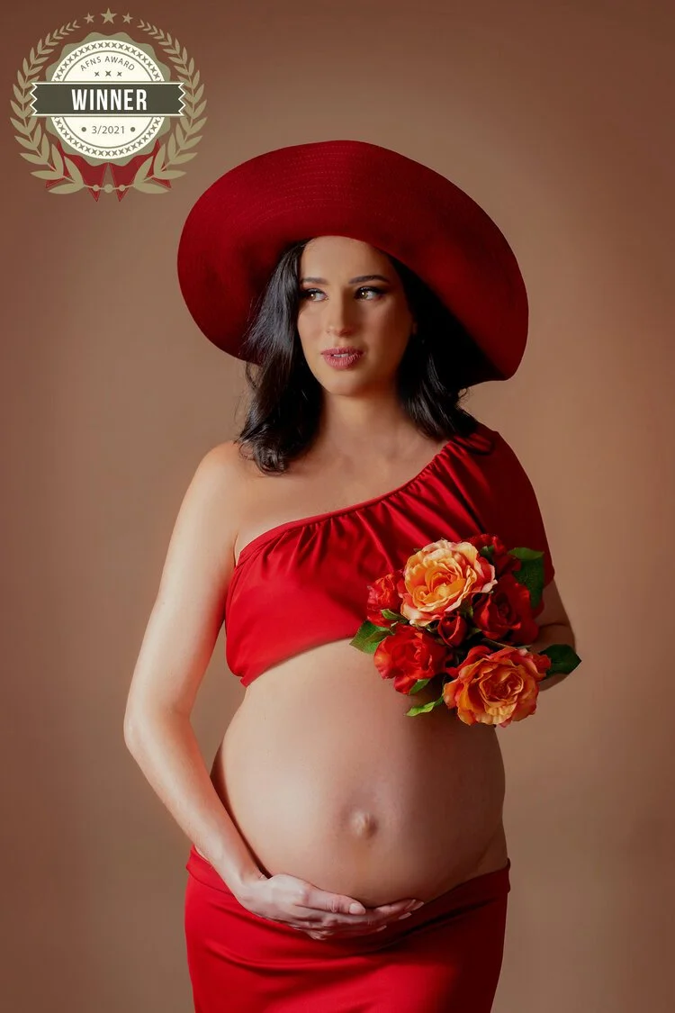 Pregnant woman in a red dress and wide-brimmed red hat holding a bouquet of orange and red flowers, standing against a plain background with a winner badge in the upper left corner.