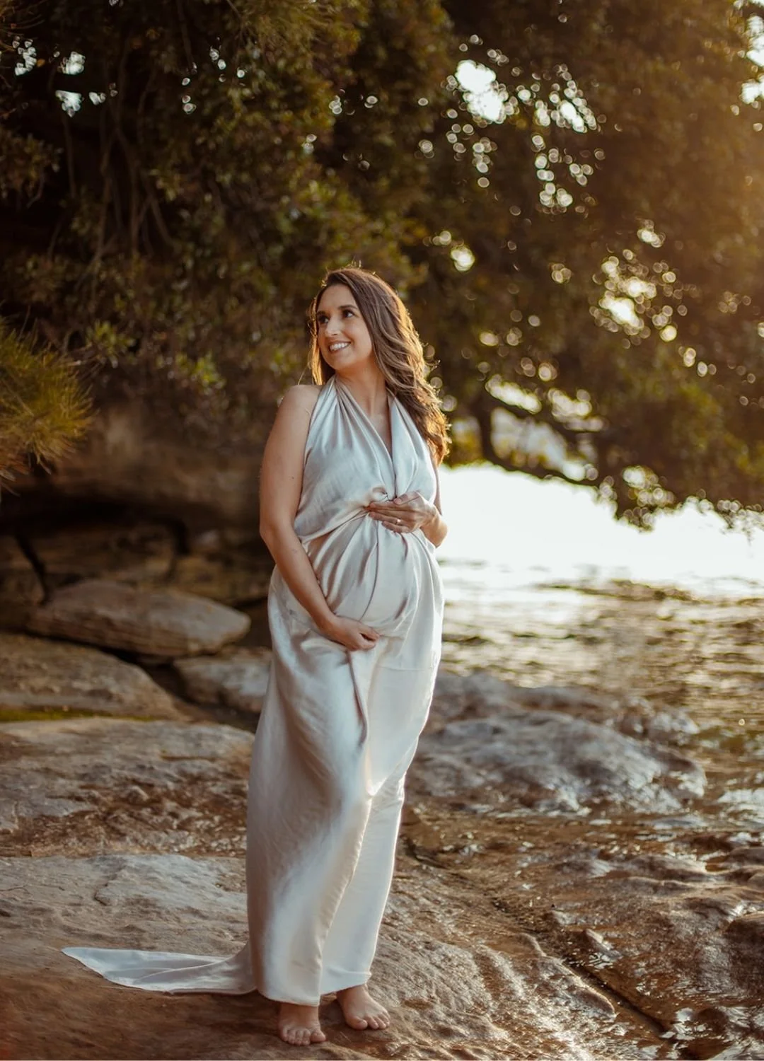 A woman wearing an elegant white dress standing barefoot on a rocky beach during sunset, with trees and water in the background.