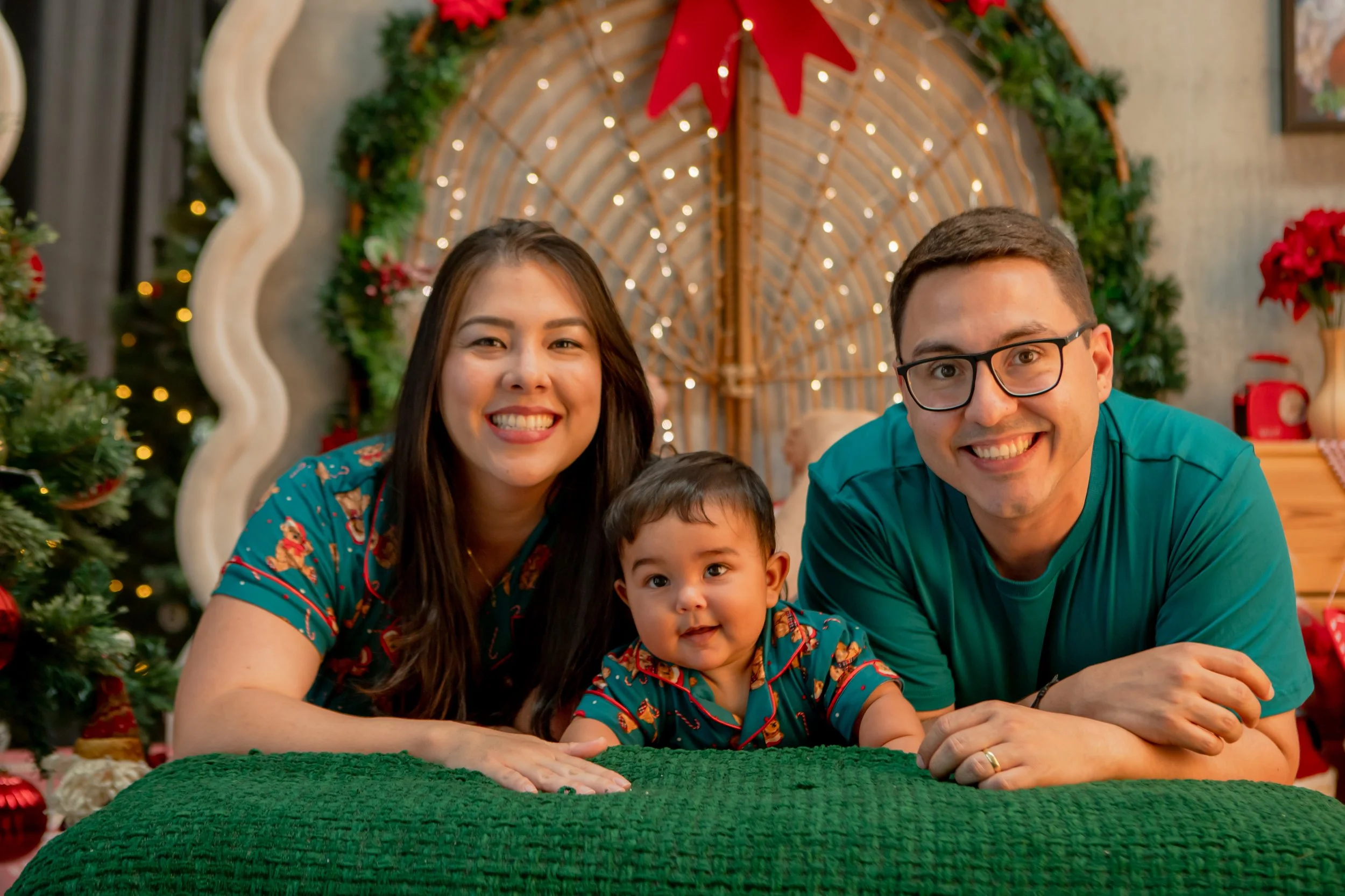 A family of three lying on their stomachs in front of Christmas decor, smiling at the camera. The woman has long dark hair, the man has short dark hair and glasses, and a young child with dark hair is between them. The background features a decorated