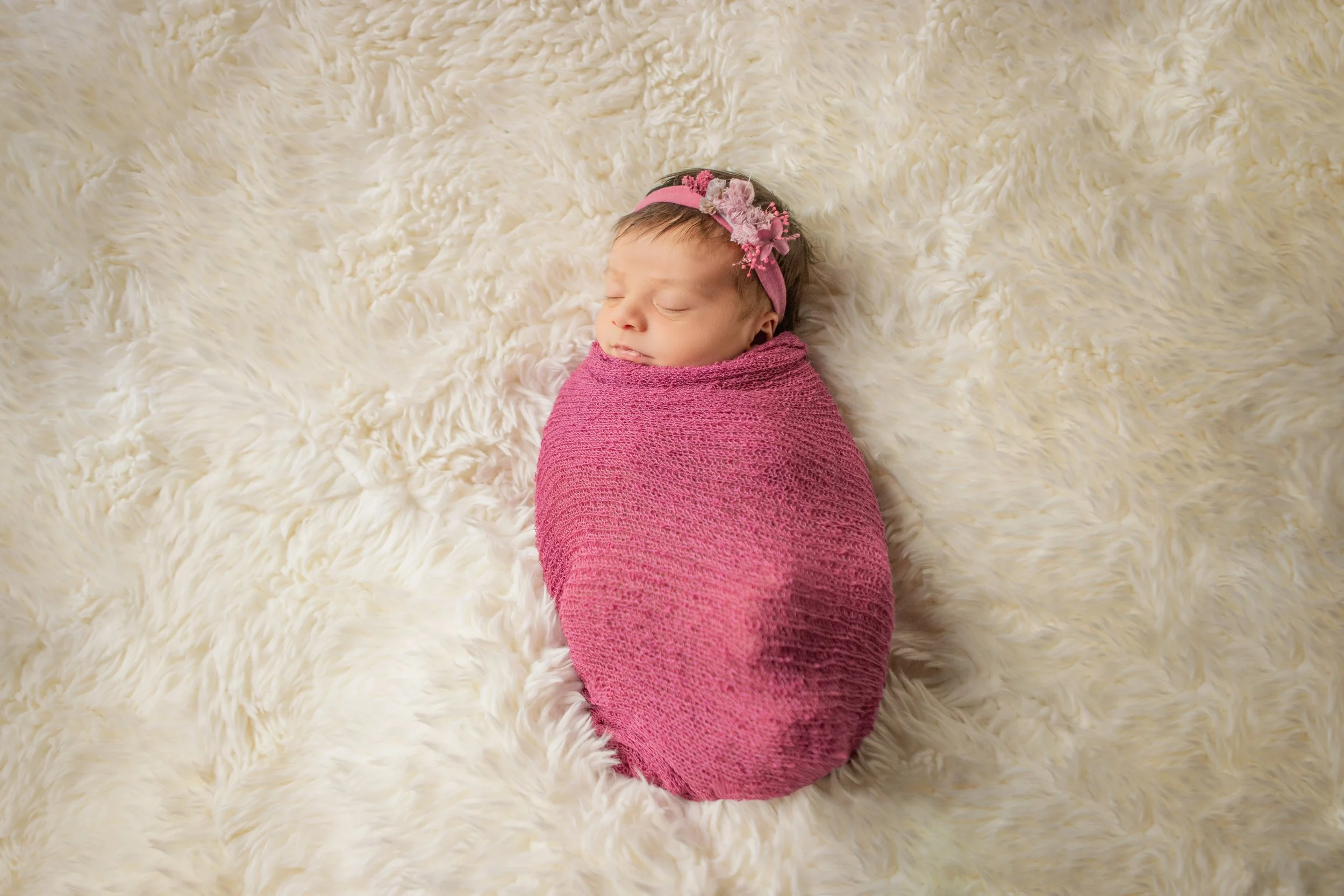 A newborn baby girl wrapped in a pink blanket, lying on a fluffy cream-colored surface, wearing a pink headband with flowers, sleeping peacefully.