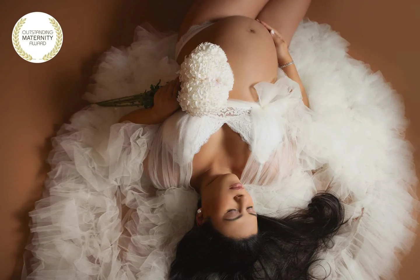 A pregnant woman in a white wedding dress lying on a soft surface, holding a bouquet of white flowers, with her eyes closed and head tilted to the side.