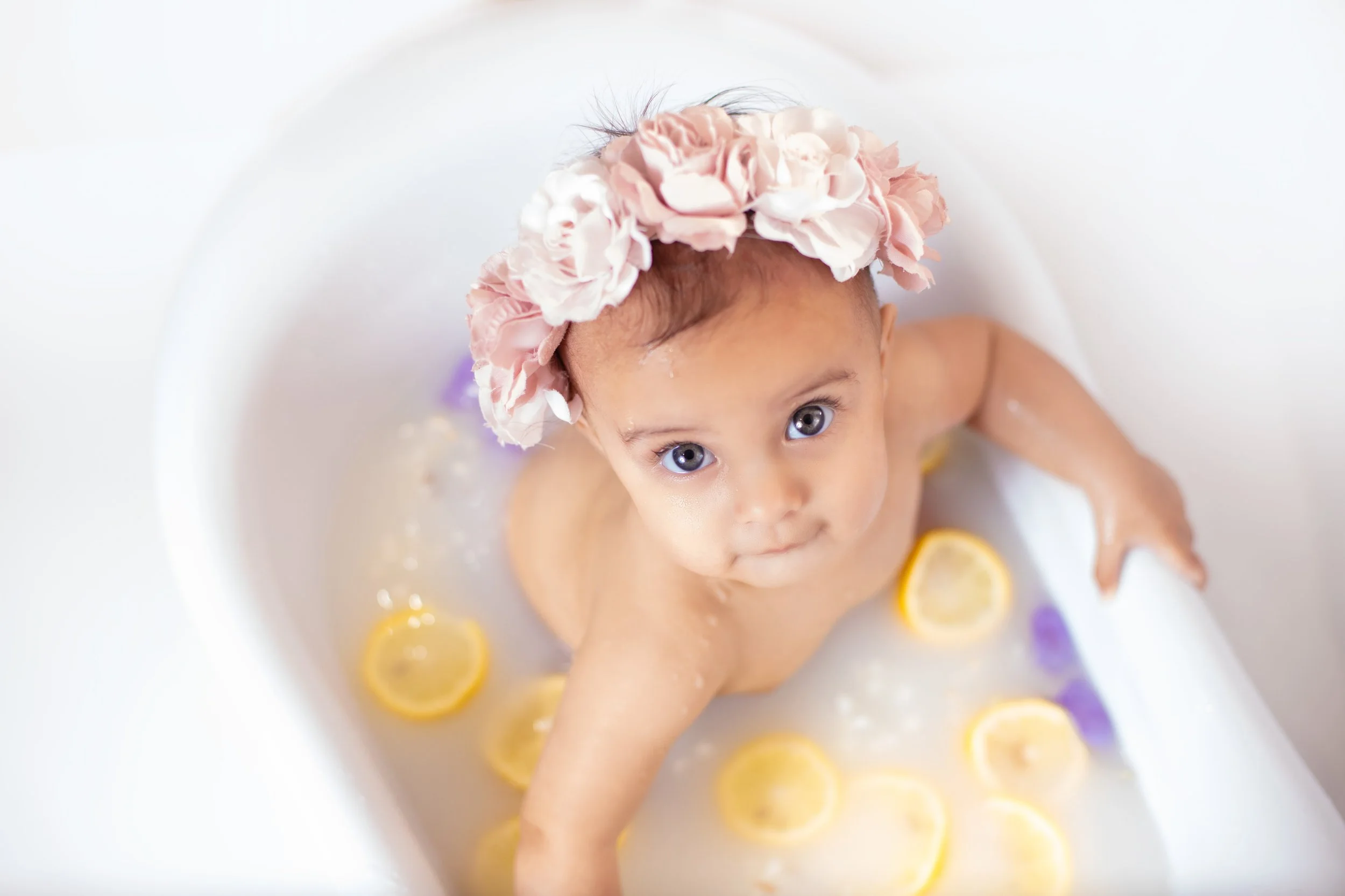 A young child taking a milk bath with lemon slices and a pink flower headband, looking up at the camera.