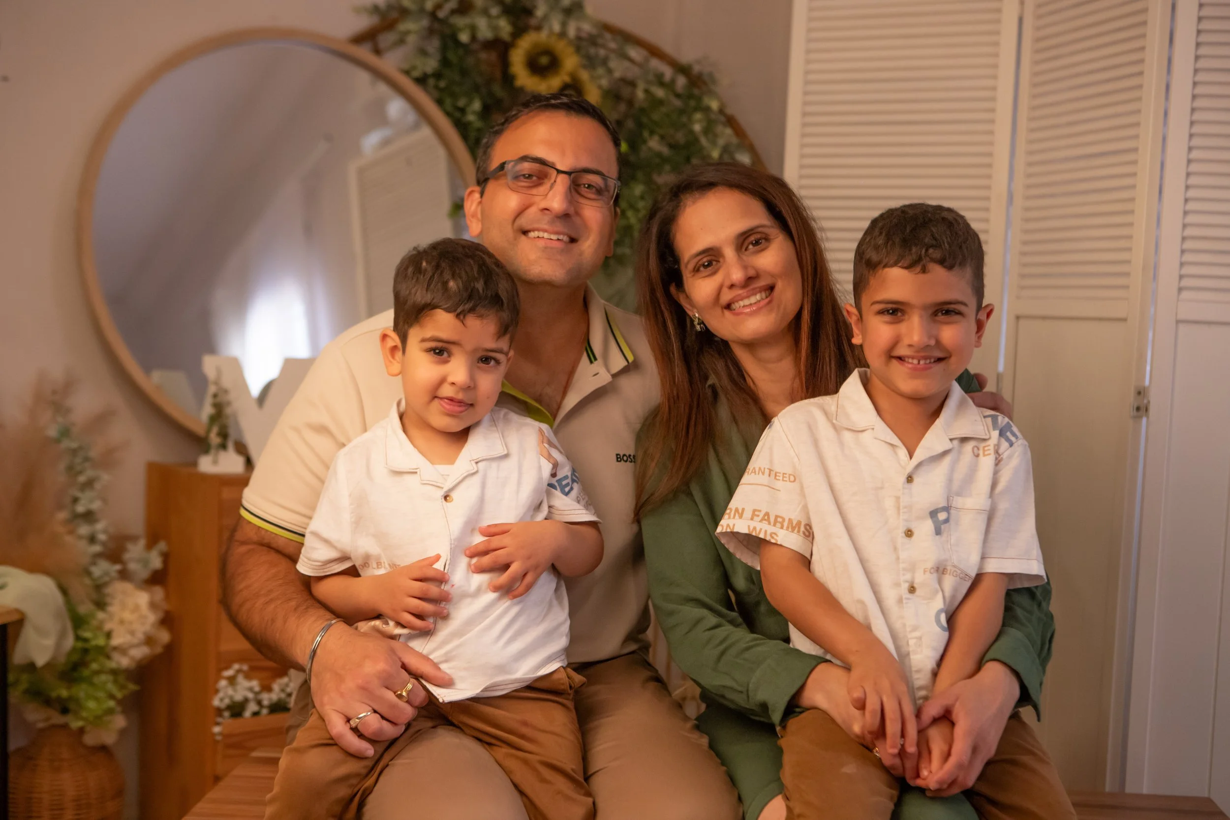 A happy family of four, with two parents and two young sons, smiling and sitting close together in a cozy, warmly lit room decorated with flowers and a round mirror in the background.