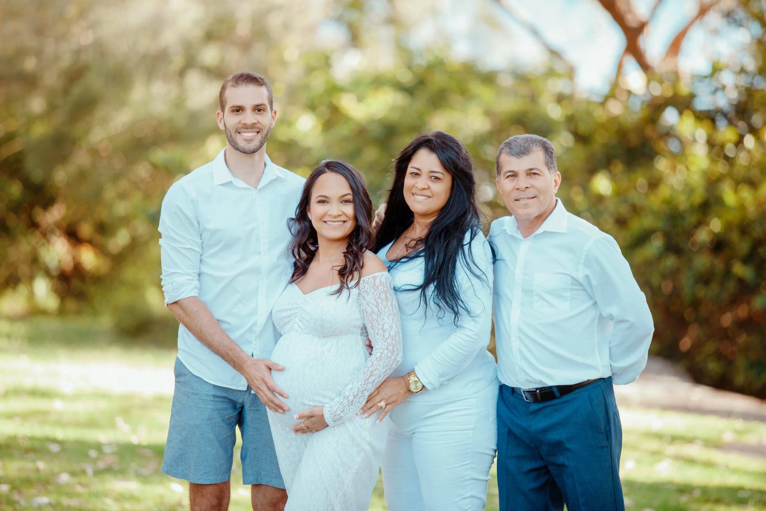 Family of four posing outdoors on a sunny day, with a pregnant woman in a white lace dress, two men, and a woman in white tops and pants, standing in front of a blurred green and brown nature background.