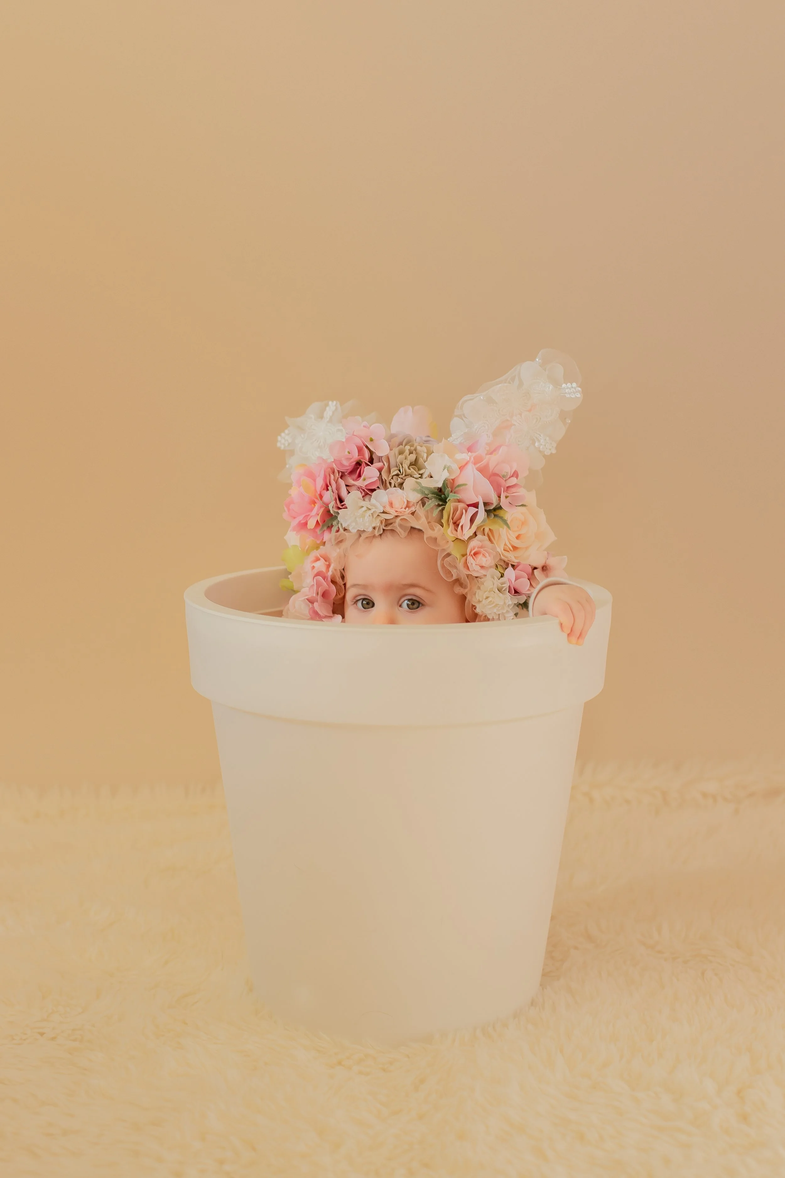A baby girl peeking out from inside a large white flowerpot, wearing a floral headband with pink, beige, and white flowers and lace bunny ears, against a soft beige background.