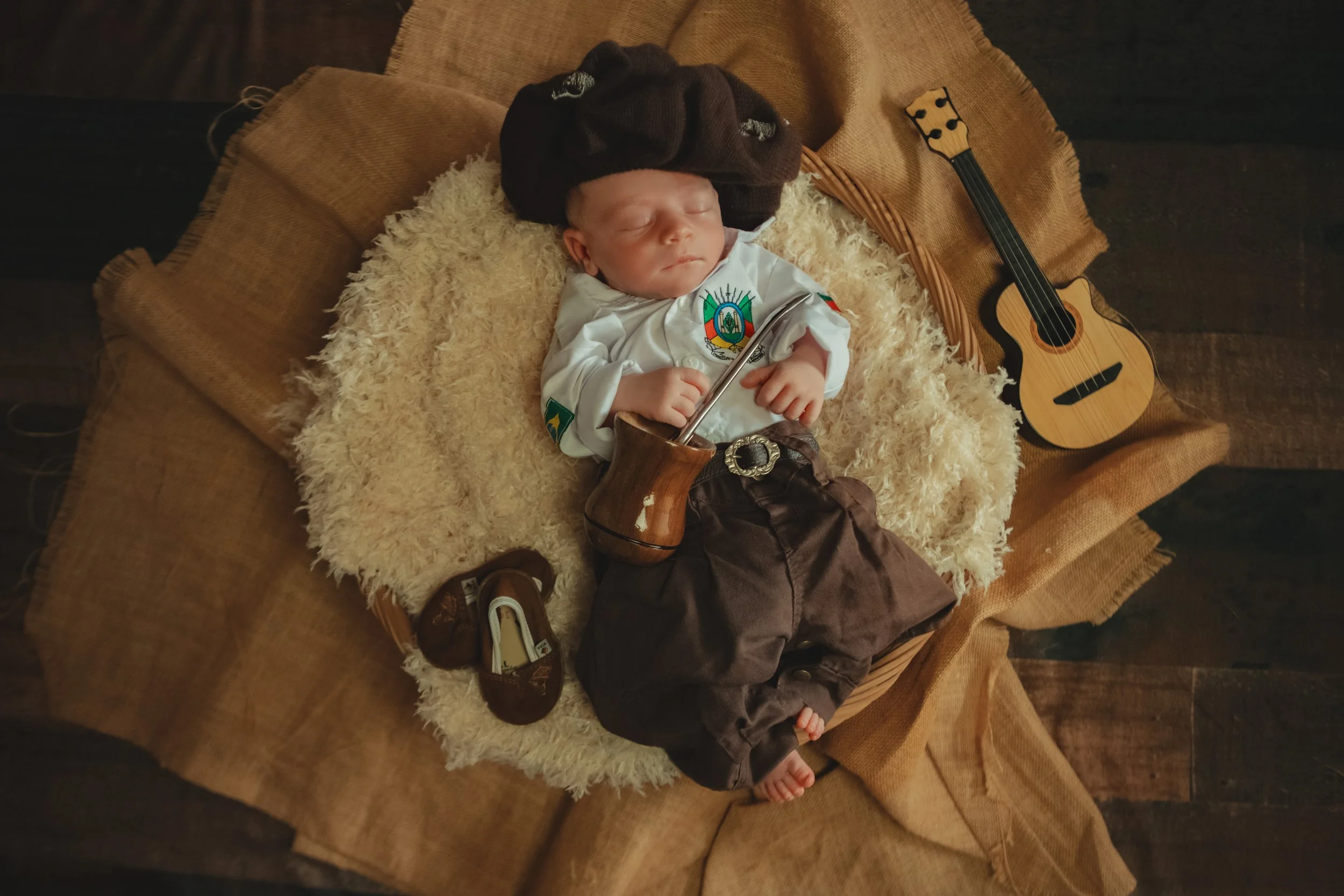 A sleeping baby boy dressed in a white shirt with colorful patches, brown pants, and a black cap lying on a fluffy cream-colored blanket inside a wicker basket. A small wooden guitar is next to him on a brown cloth-covered surface, and a pair of dark
