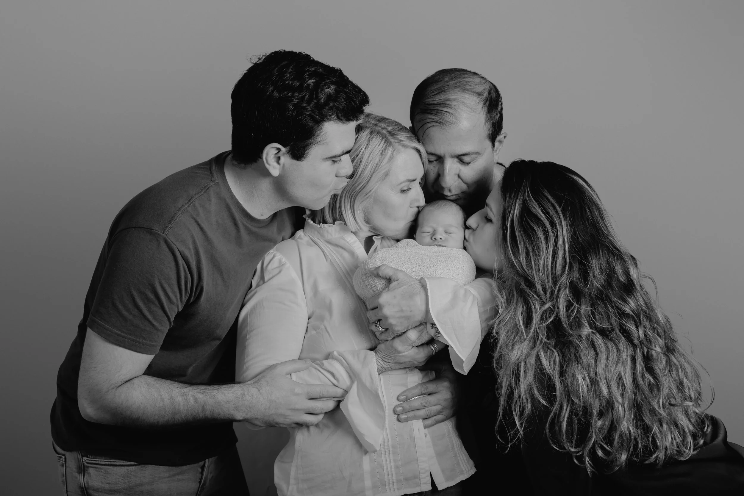 A black and white photograph of a family gathered around a newborn, kissing, and holding the baby, with two adults and a young woman in close proximity.